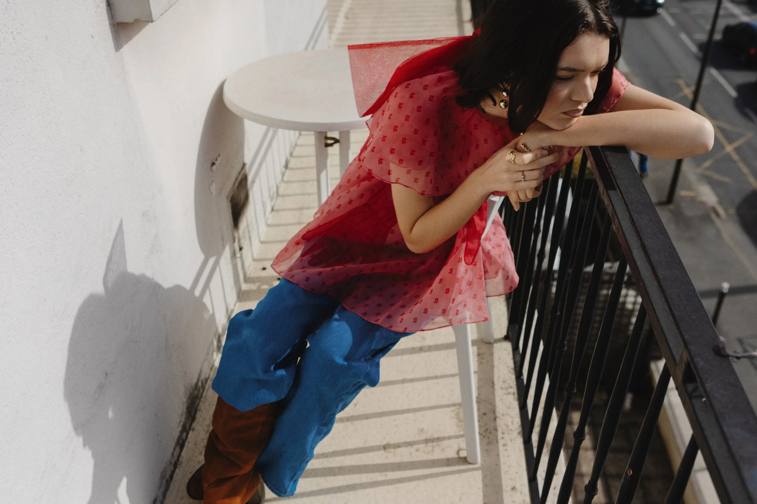 A woman with dark hair and earrings, wearing a pink floral blouse, blue jeans, and brown boots, leaning on a black railing and looking down, standing on a balcony at night.