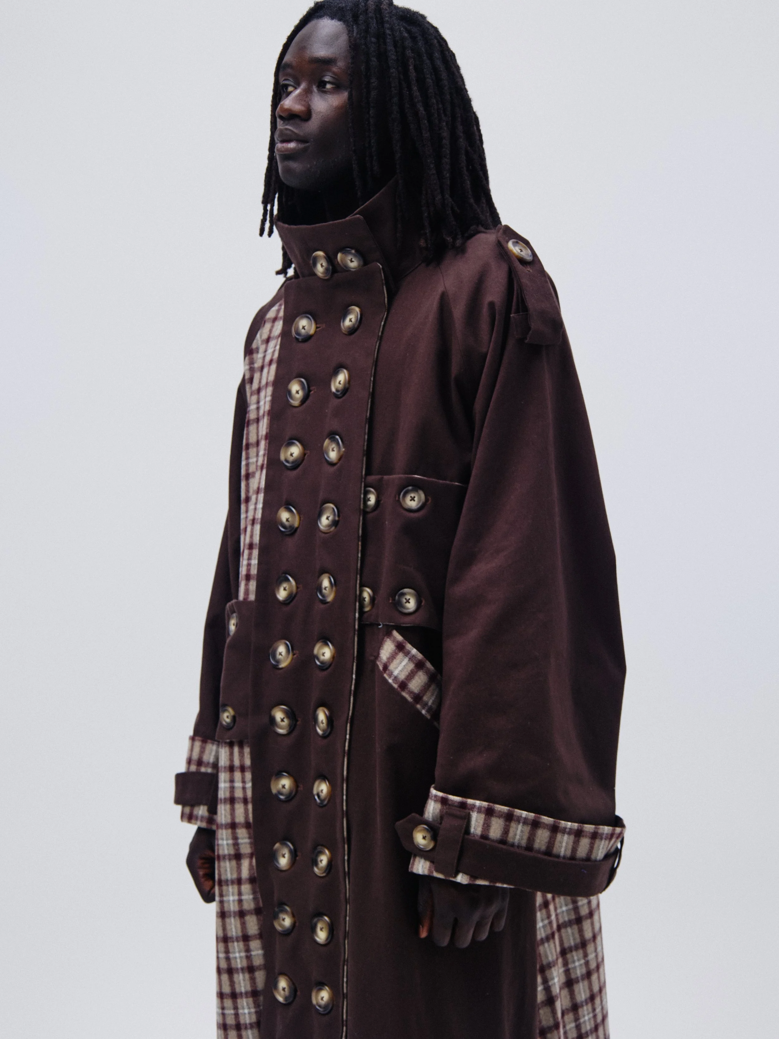 A person with long dreadlocks wearing a brown trench coat with large buttons and plaid details on the collar and cuffs, standing against a plain light gray background.
