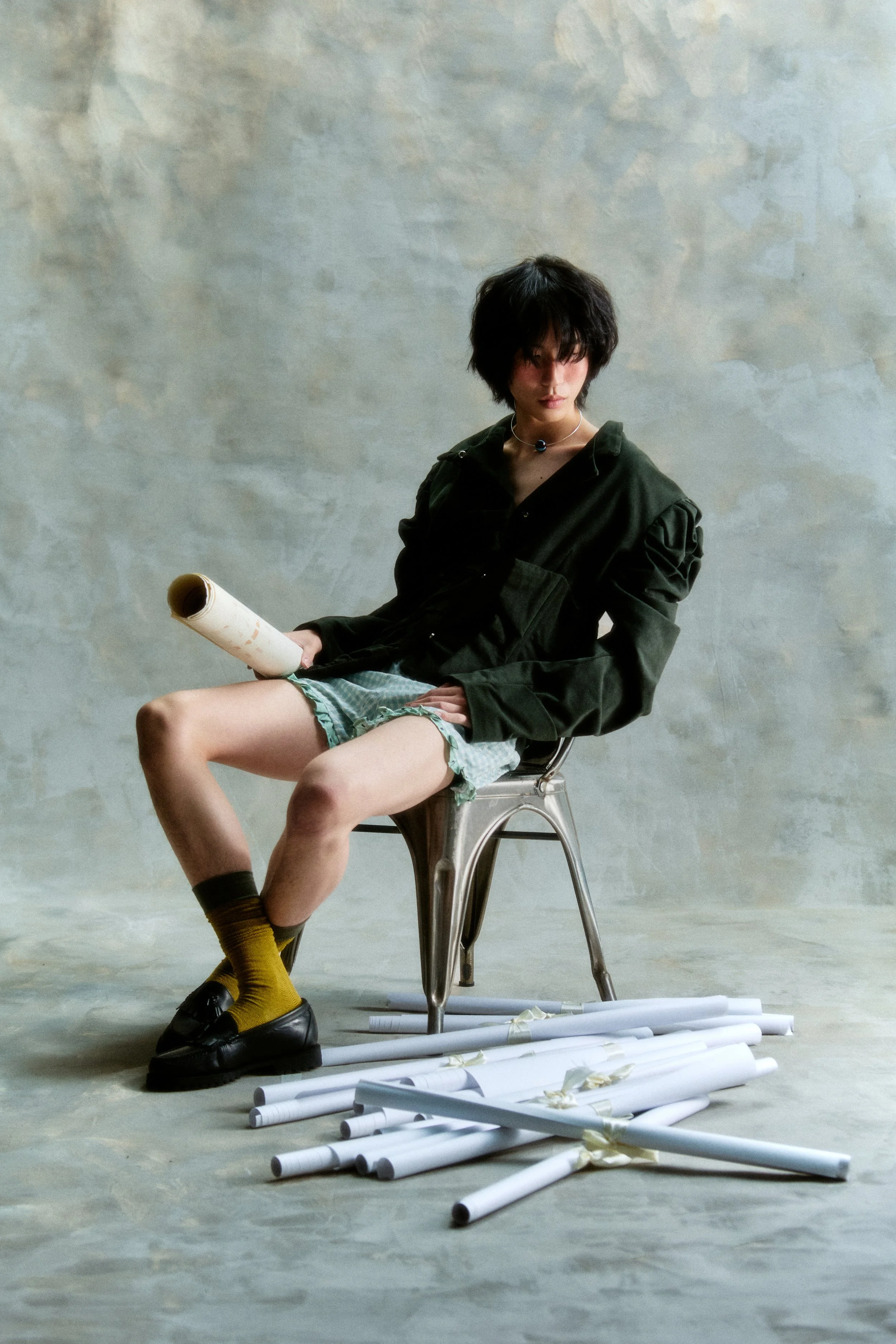 A person with short dark hair wearing a black jacket, patterned shorts, yellow socks, and black shoes, sitting on a metal chair in front of a plain textured wall. They are holding a rolled-up paper in one hand and are surrounded by scattered rolled-u