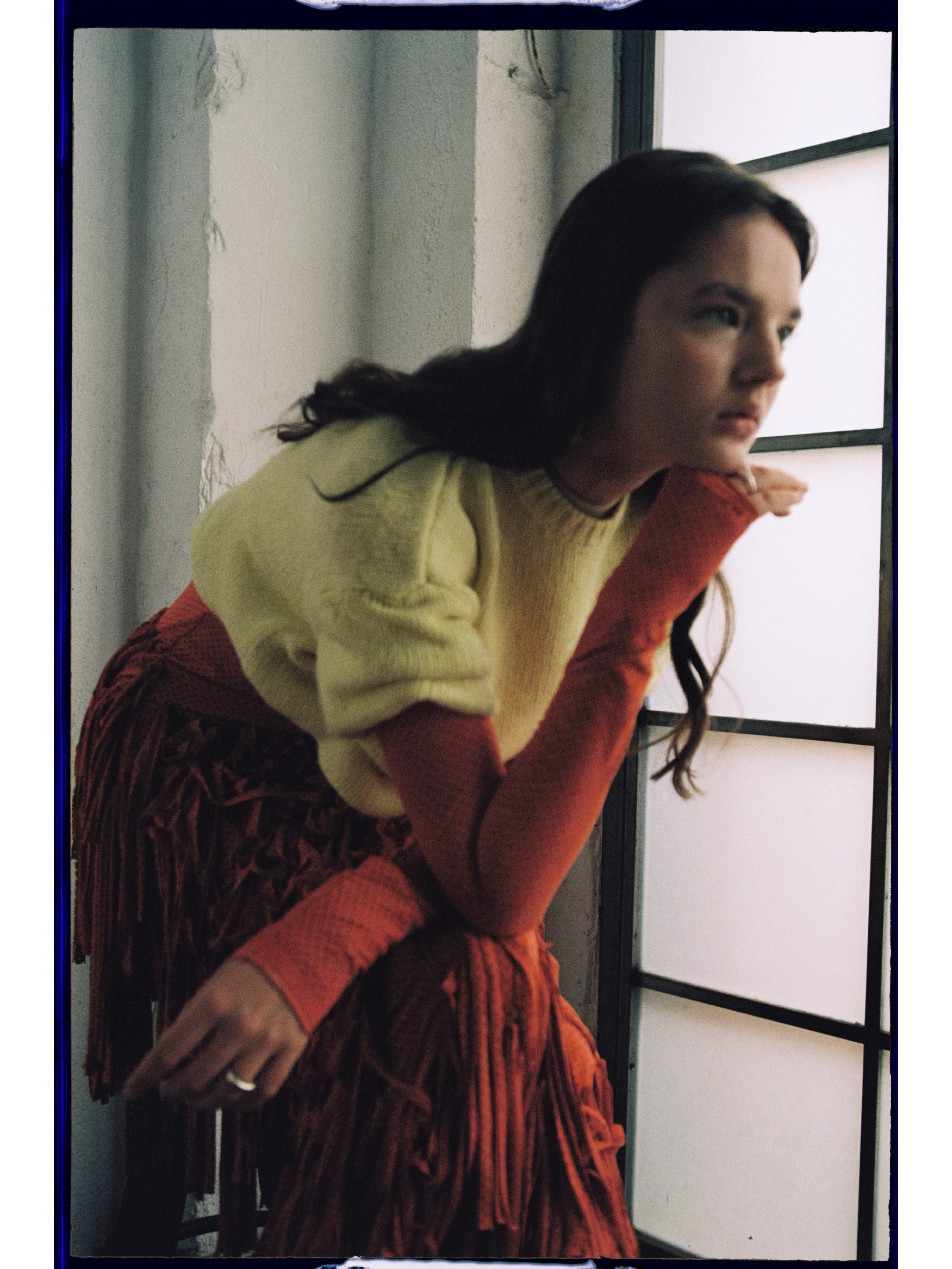 A woman with long dark hair wearing a beige top and red patterned skirt, leaning against a wall near a window, looking to the side.