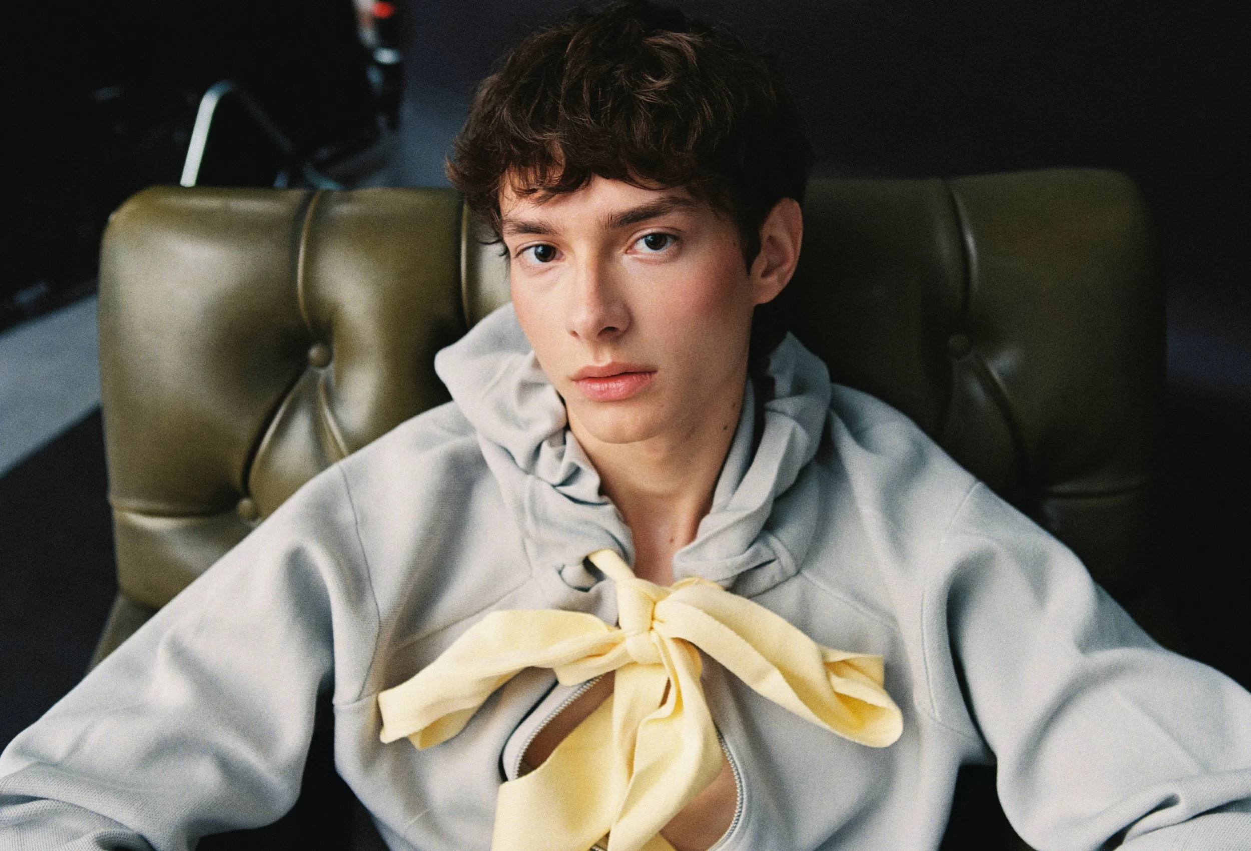 A young person with short curly hair and light skin sitting on a green leather chair, wearing a gray hoodie and a yellow scarf, looking directly at the camera.