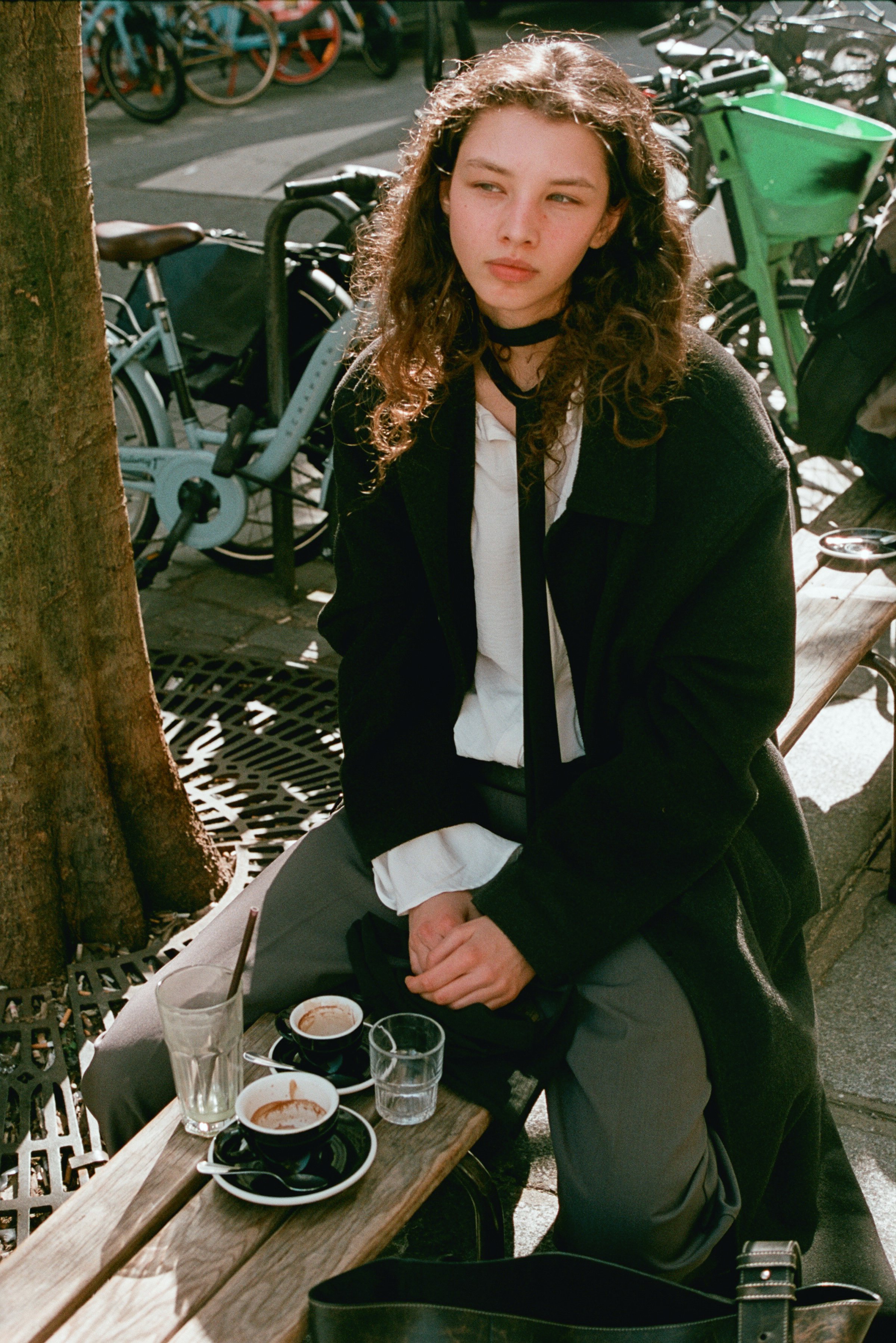 A young woman with long curly hair sitting outdoors at a café table with multiple empty dishes and glasses, some with remnants of drinks, during daytime.