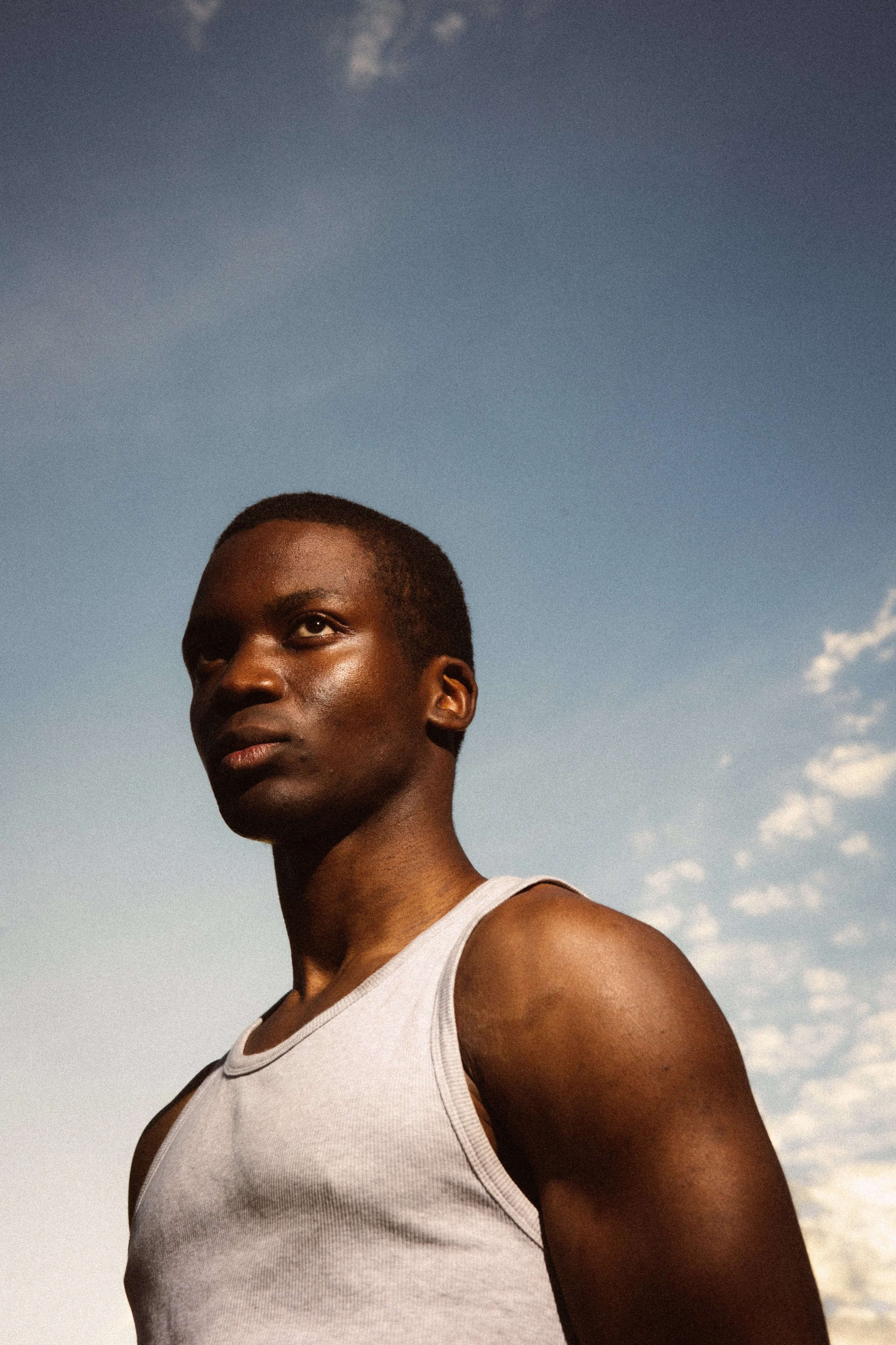 Close-up of a person with dark skin wearing a white tank top, looking to the side, against a sky background with clouds and a faint moon.