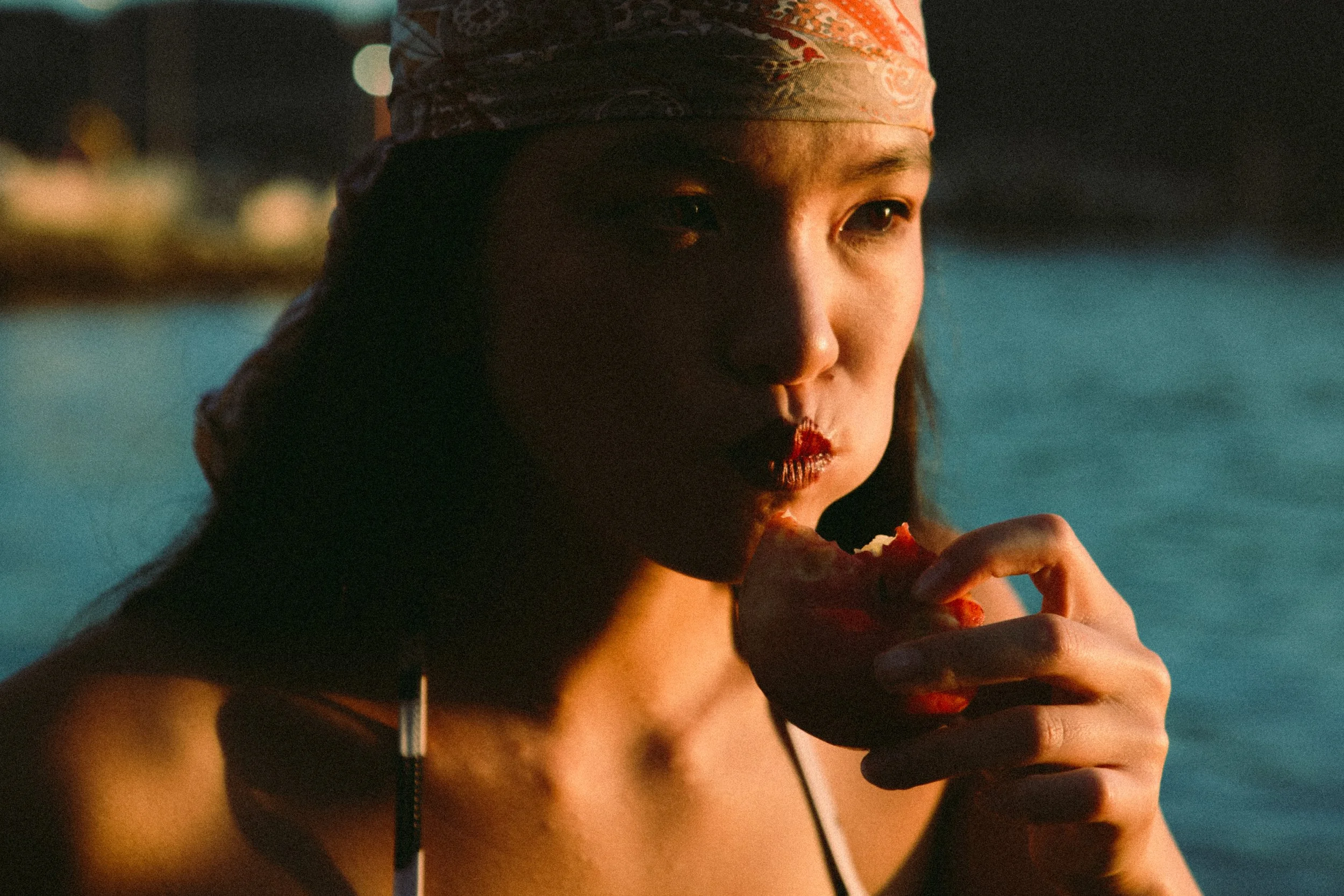 Close-up of a woman with a colorful headscarf eating a slice of pizza at night near a body of water.