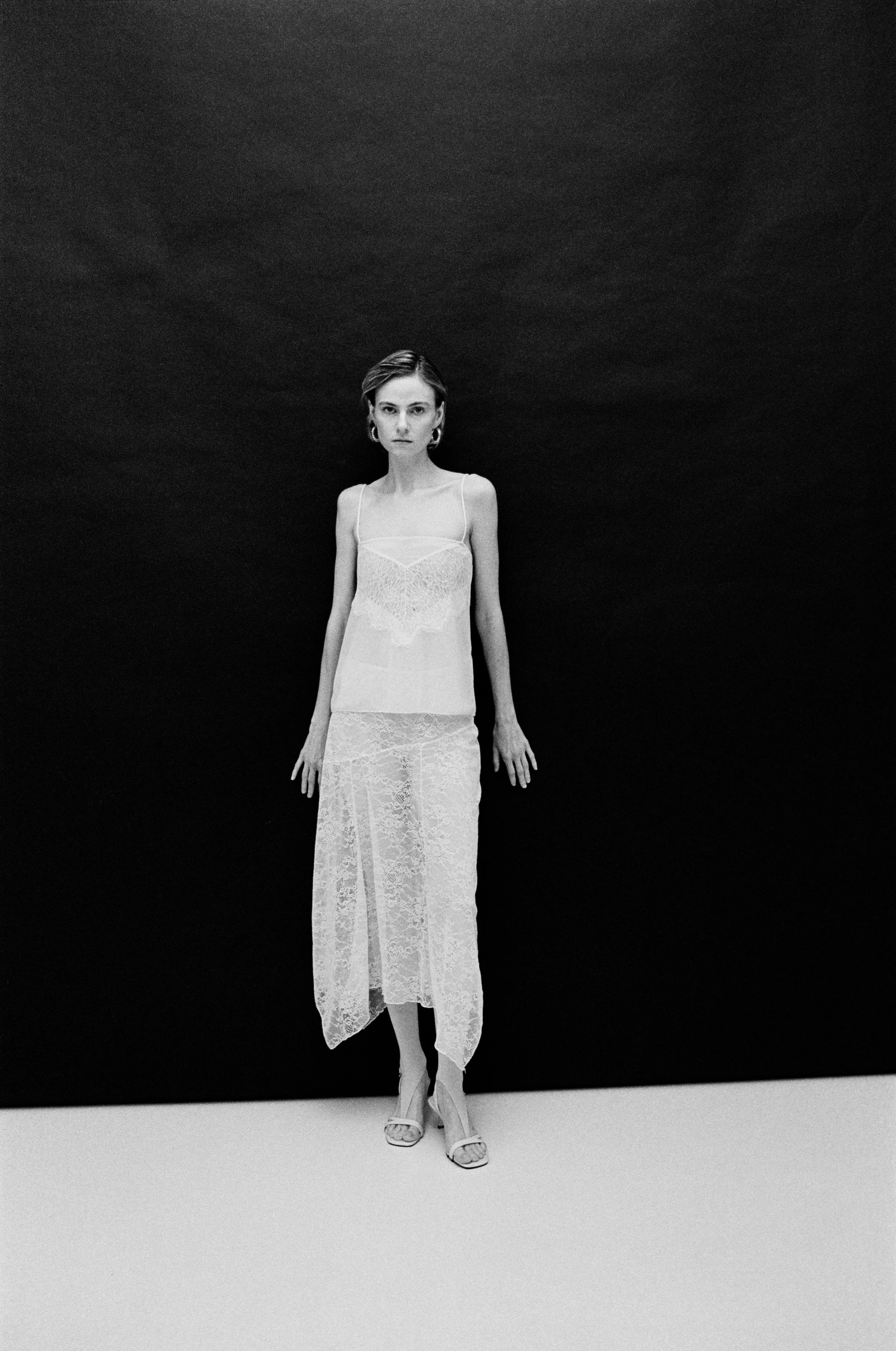 A woman wearing a sleeveless lace dress and high heels standing and posing in front of a dark background.