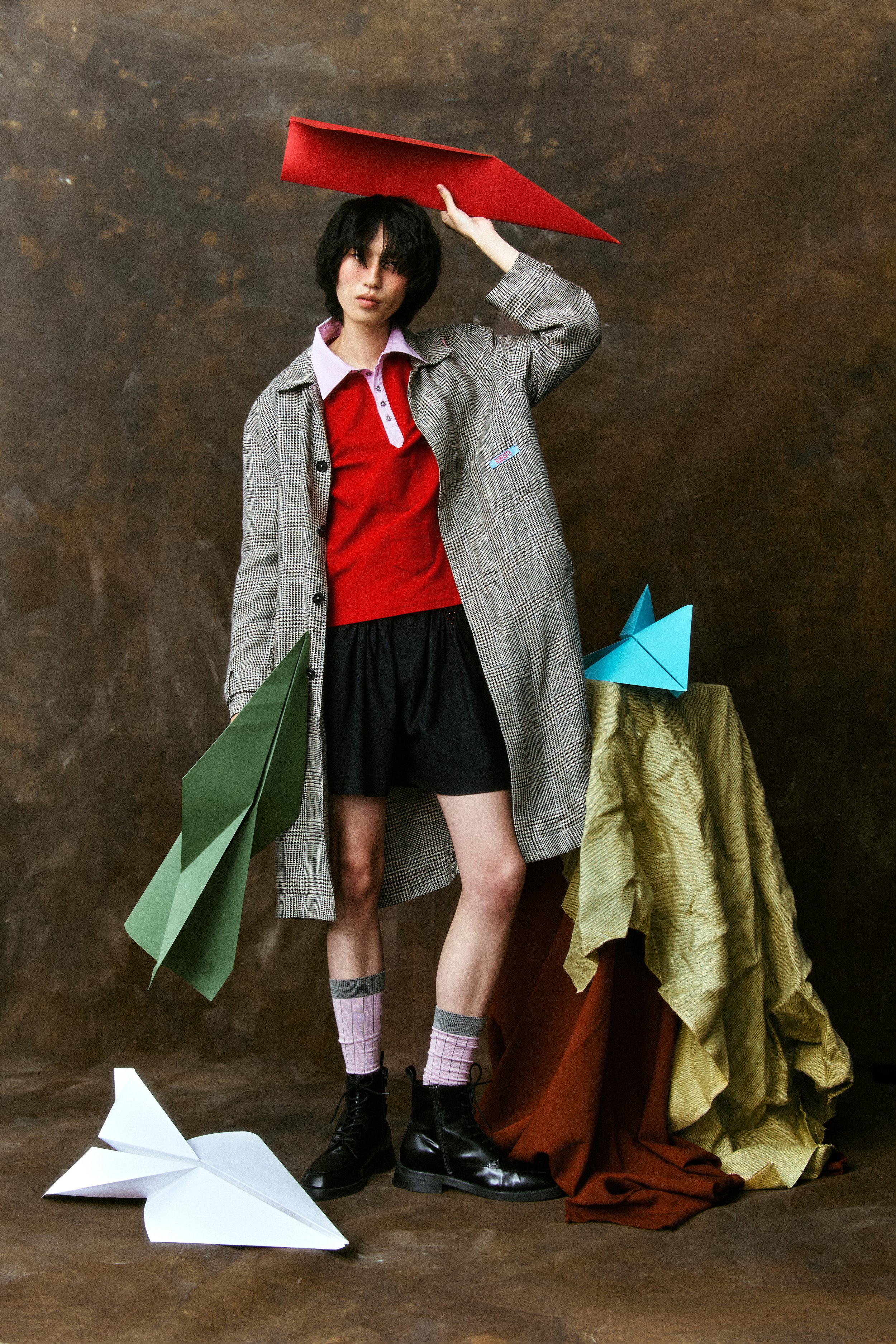 A young woman with short black hair wearing a checkered coat, red top, and shorts standing against a brown background. She is holding a large red paper airplane above her head. There are paper airplanes on the floor and on a table beside her, with va
