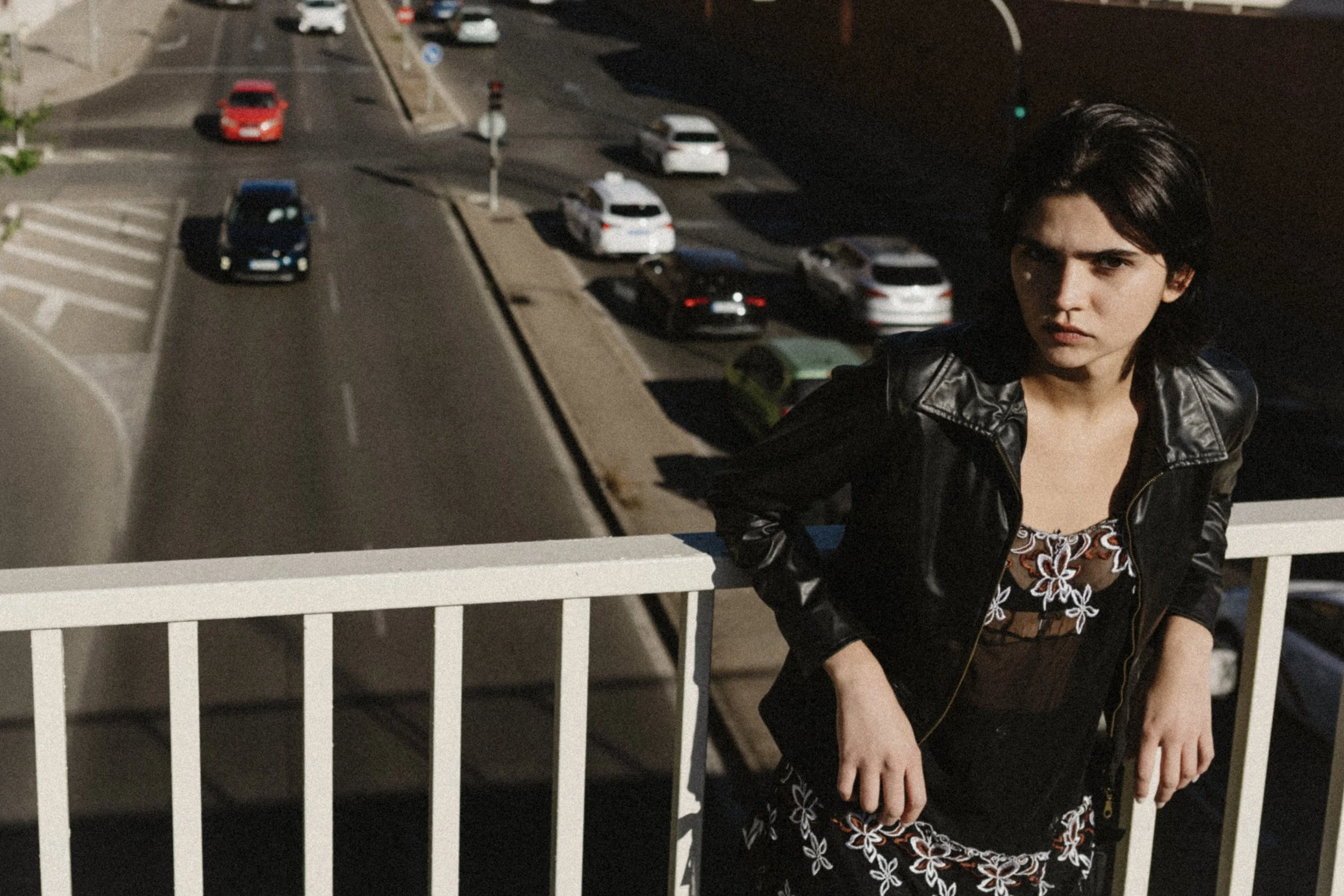 A woman wearing a black leather jacket and a black floral dress with orange and white flowers is sitting on a white fence at night. She has short dark hair and is looking to her right.