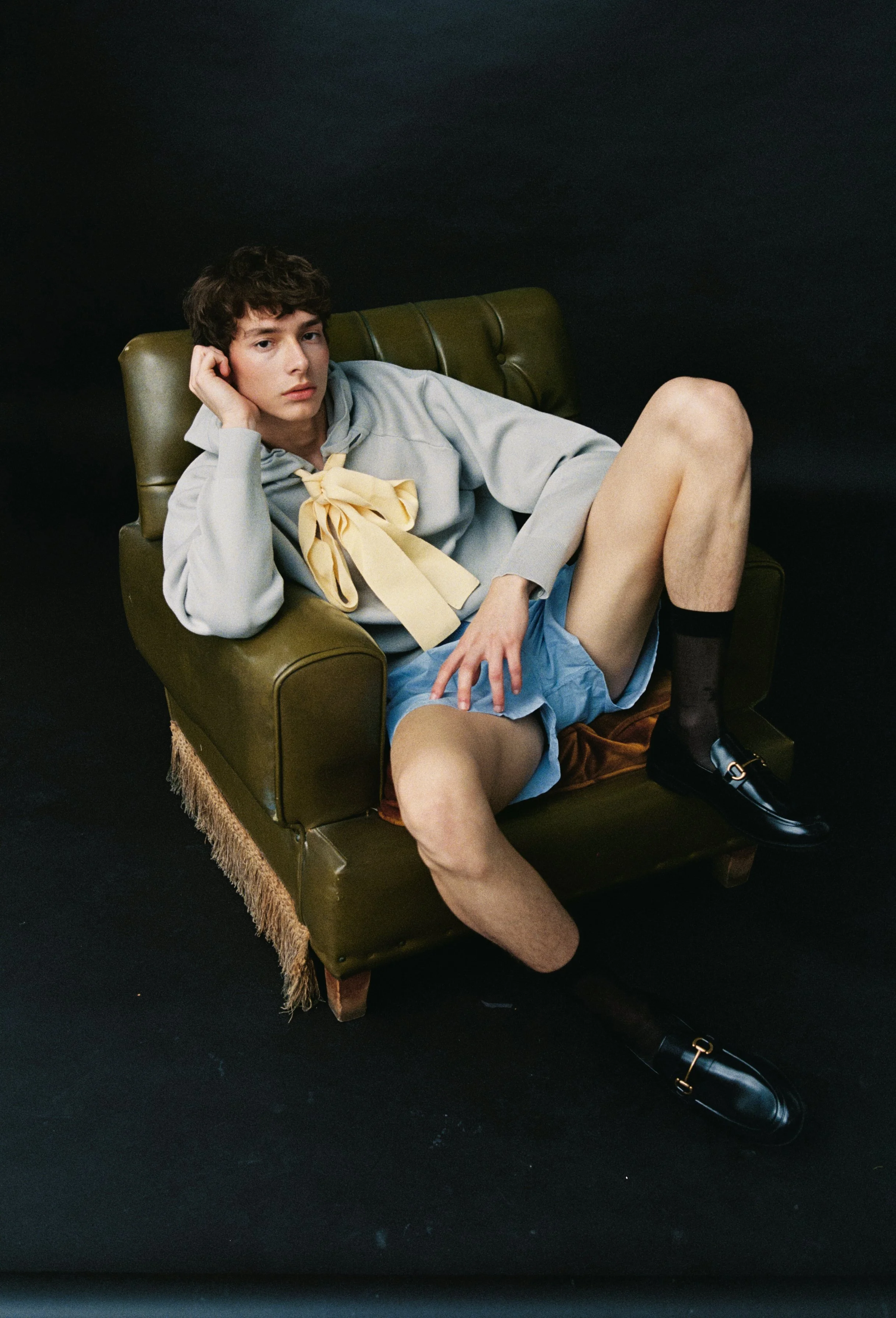 A young person with short dark hair laying on a green vintage armchair with one arm resting on the armrest and the other hand on their face. They are wearing a light gray hoodie with a yellow bow at the neck, blue shorts, and black shoes with socks. 
