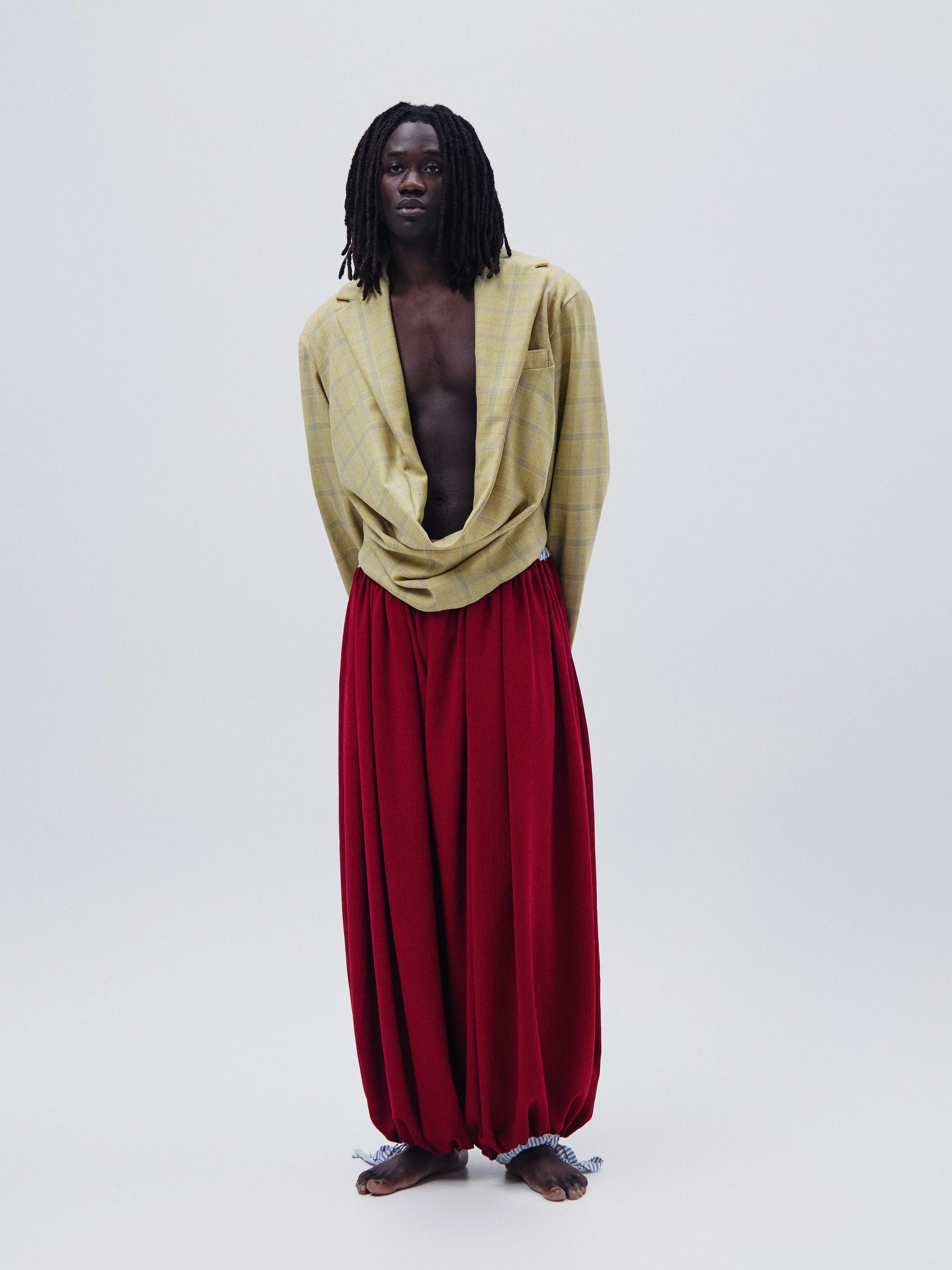 A person with dark skin and long dreadlocks standing barefoot against a plain white background, wearing a yellow plaid blazer and red loose-fitting pants with striped ties at the ankles.