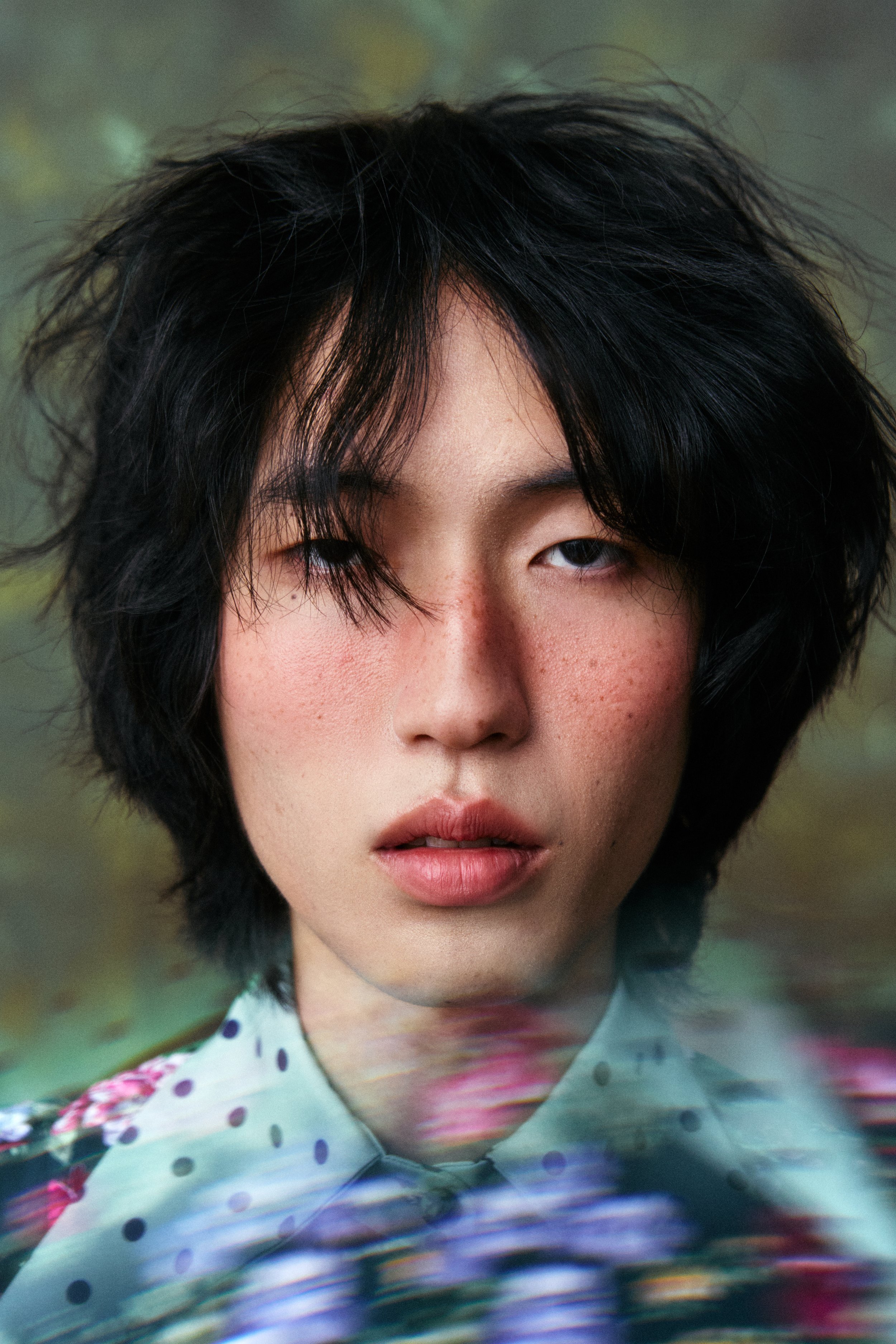 Close-up of a person's face with short, wavy black hair, partially covering one eye, and wearing a colorful polka dot shirt.