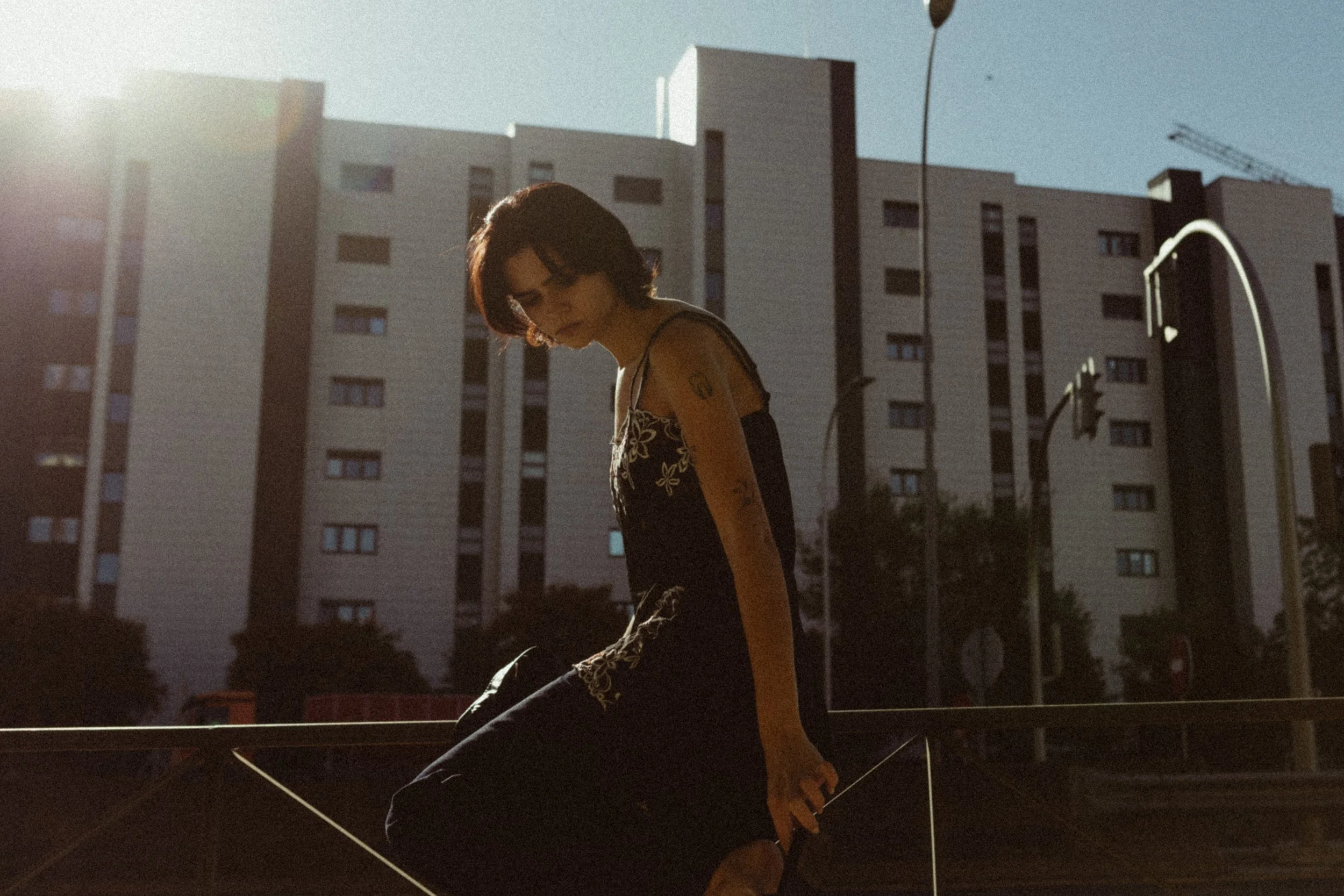 A woman with dark hair and tattoos, wearing a black dress with floral embroidery, appears to be sitting outdoors during sunset, with tall apartment buildings and streetlights in the background.