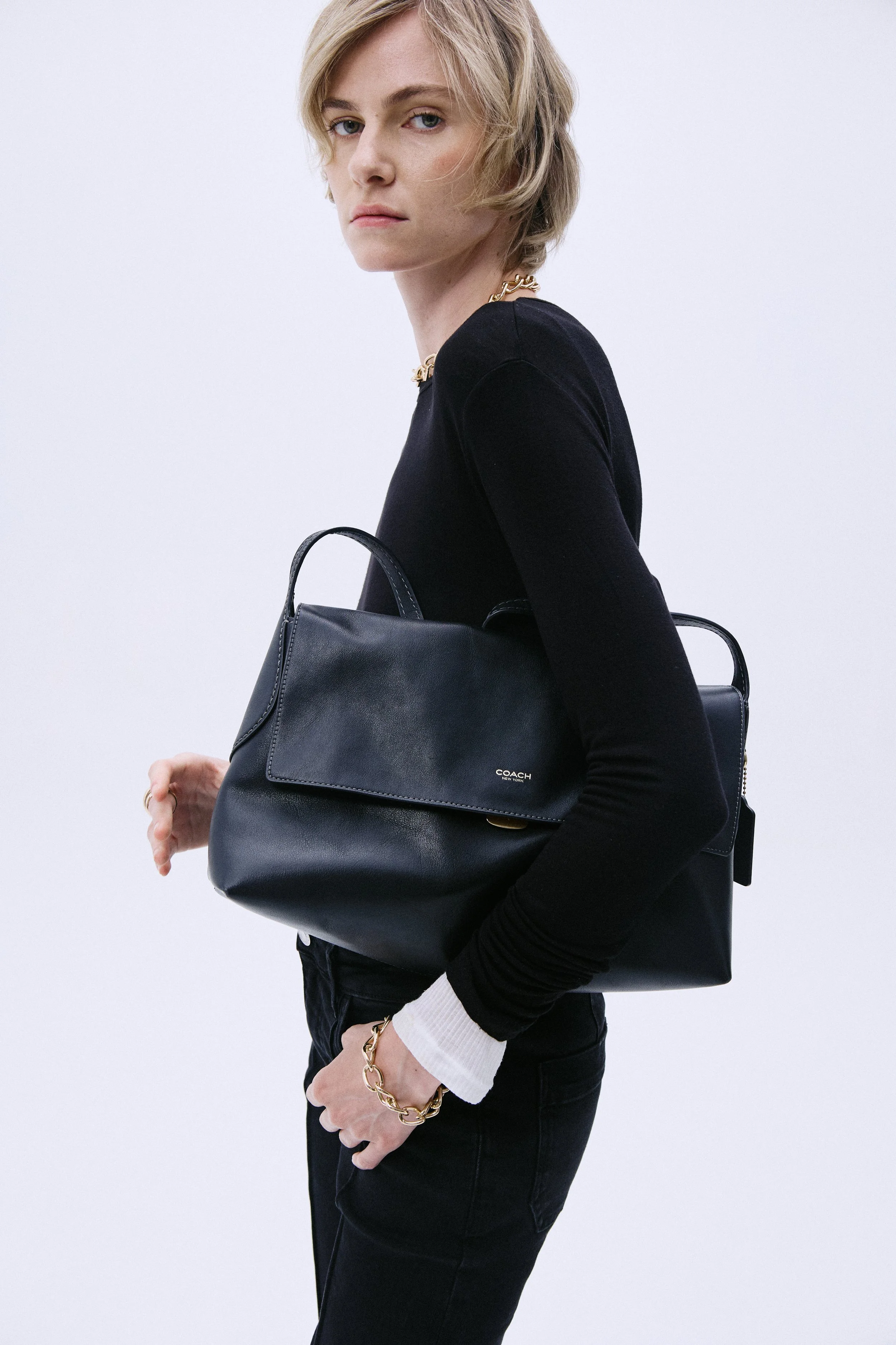 A person with short blonde hair wearing a black long-sleeve top, holding a black leather Coach handbag, dressed in black pants, and wearing a gold chain bracelet and necklace.