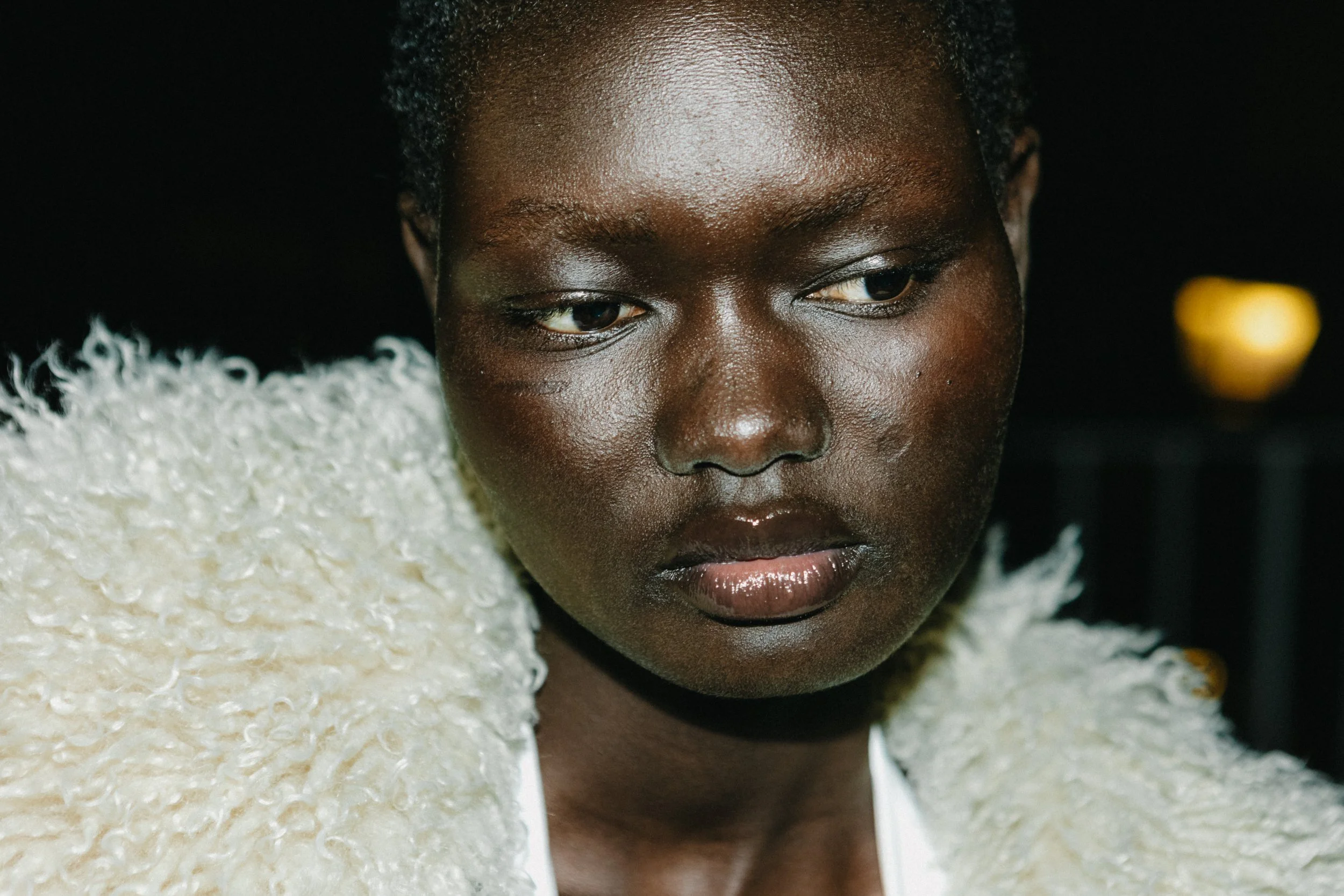 A close-up of a young woman with dark skin and short, curly hair, wearing a textured beige and white jacket, looking downward with a neutral expression.