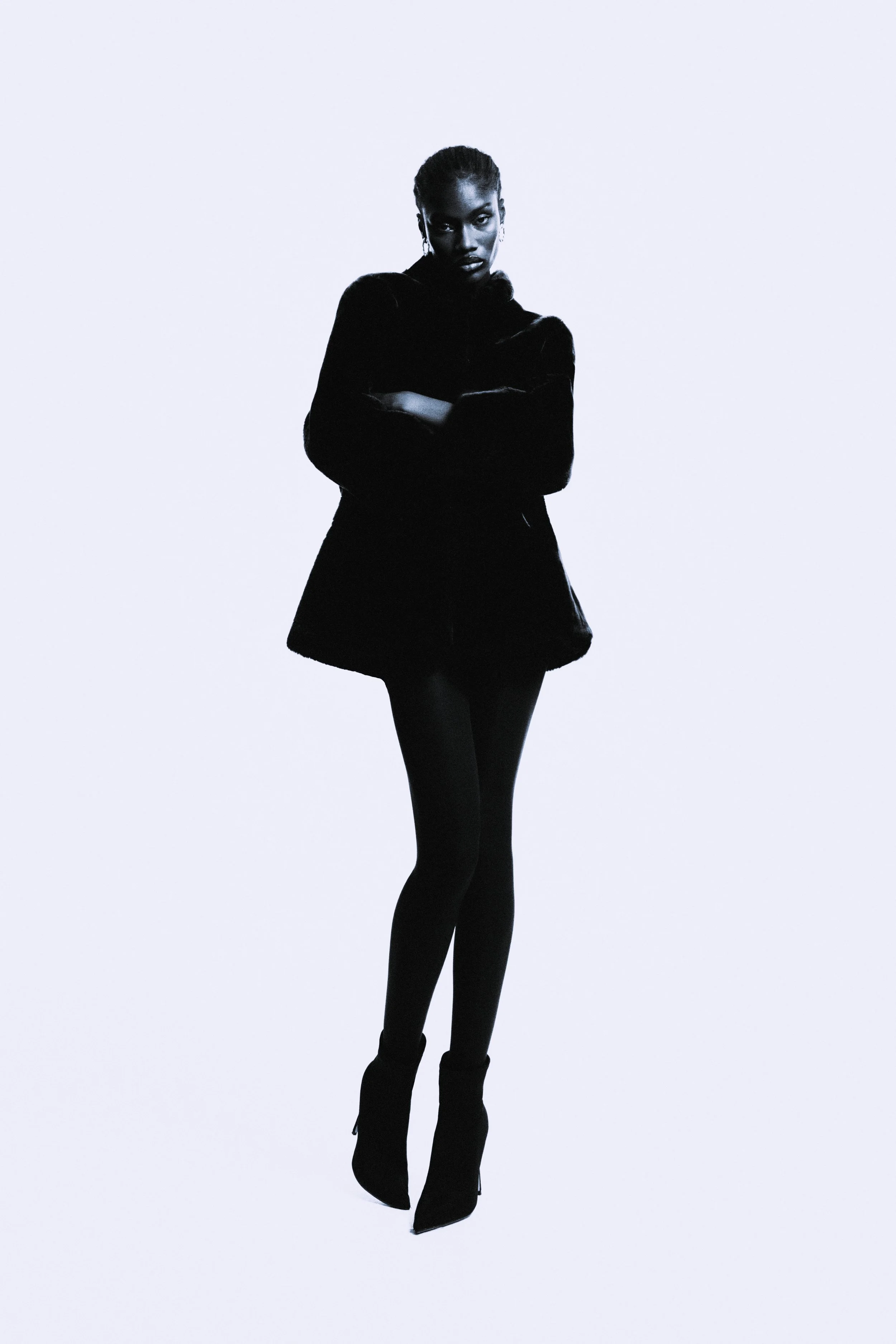 A woman with dark skin and braided hair posing against a plain light background. She is wearing a black coat, black tights, and black boots, with her arms crossed and a serious expression.