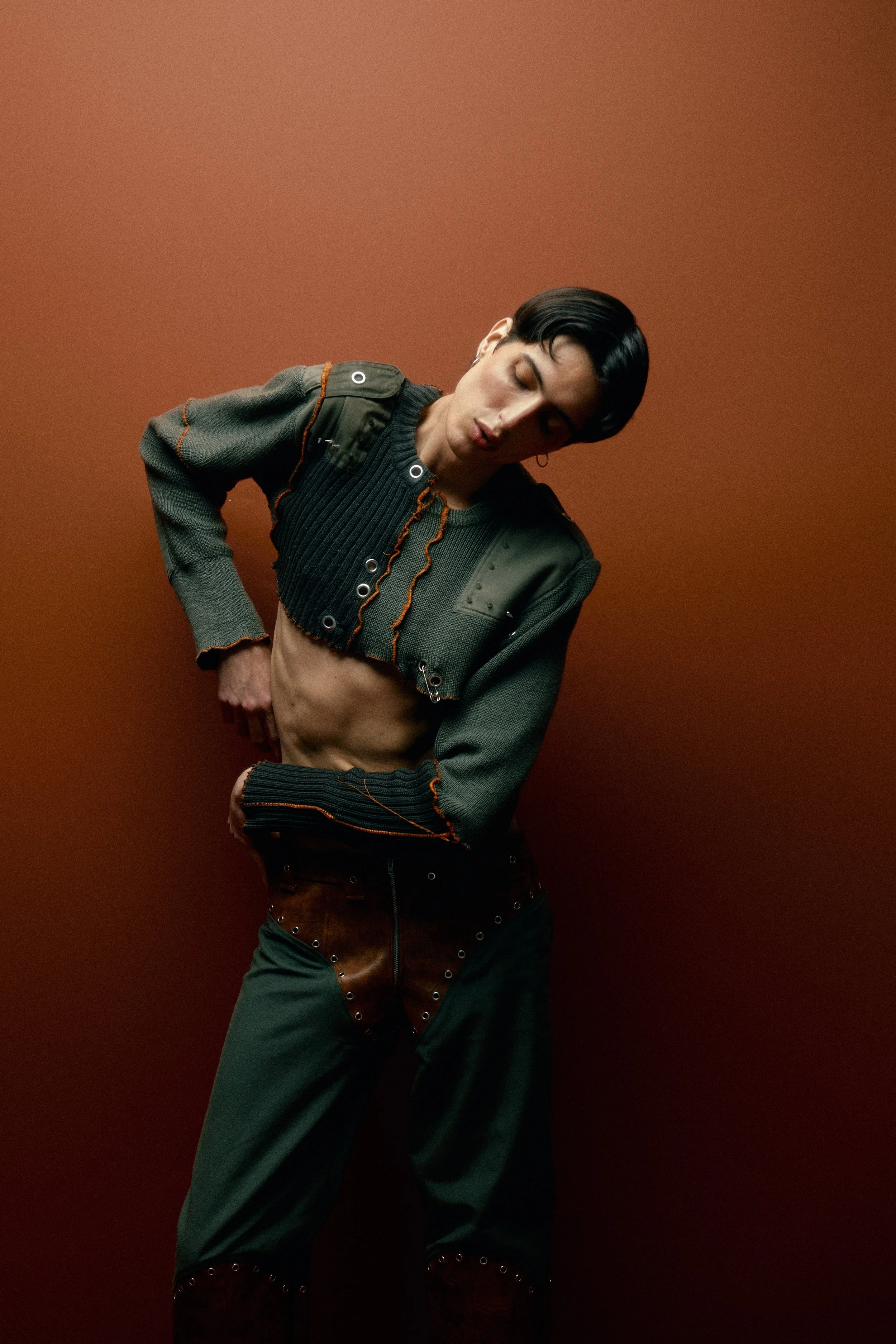A person with short black hair wearing a dark green and black jacket, cropped pants, and brown leather pants standing against a solid reddish-brown background in a fashion pose.