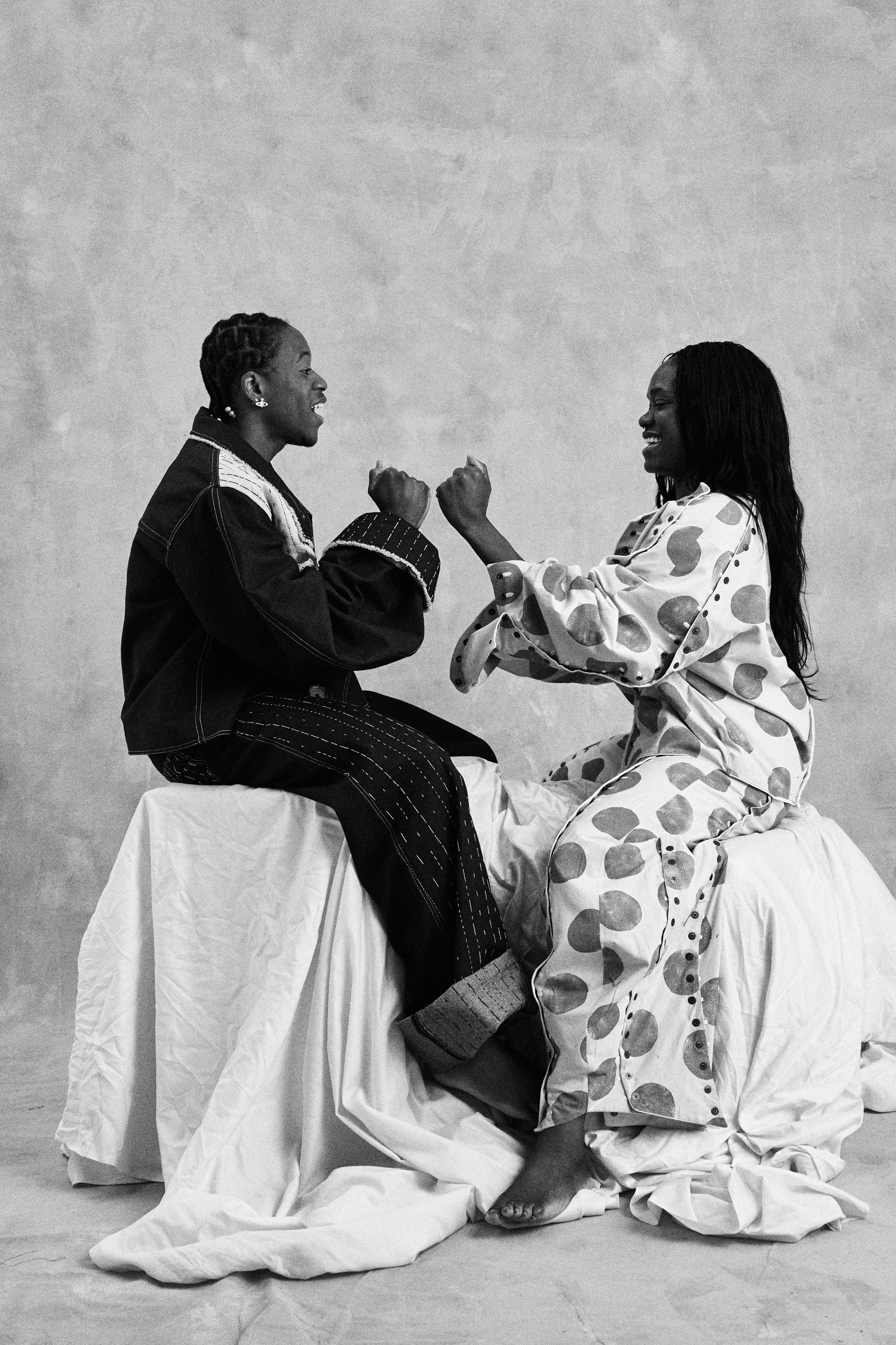 Two women facing each other with fists raised, smiling, seated on a makeshift platform with a draped cloth, wearing contrasting clothing, in a staged photo.