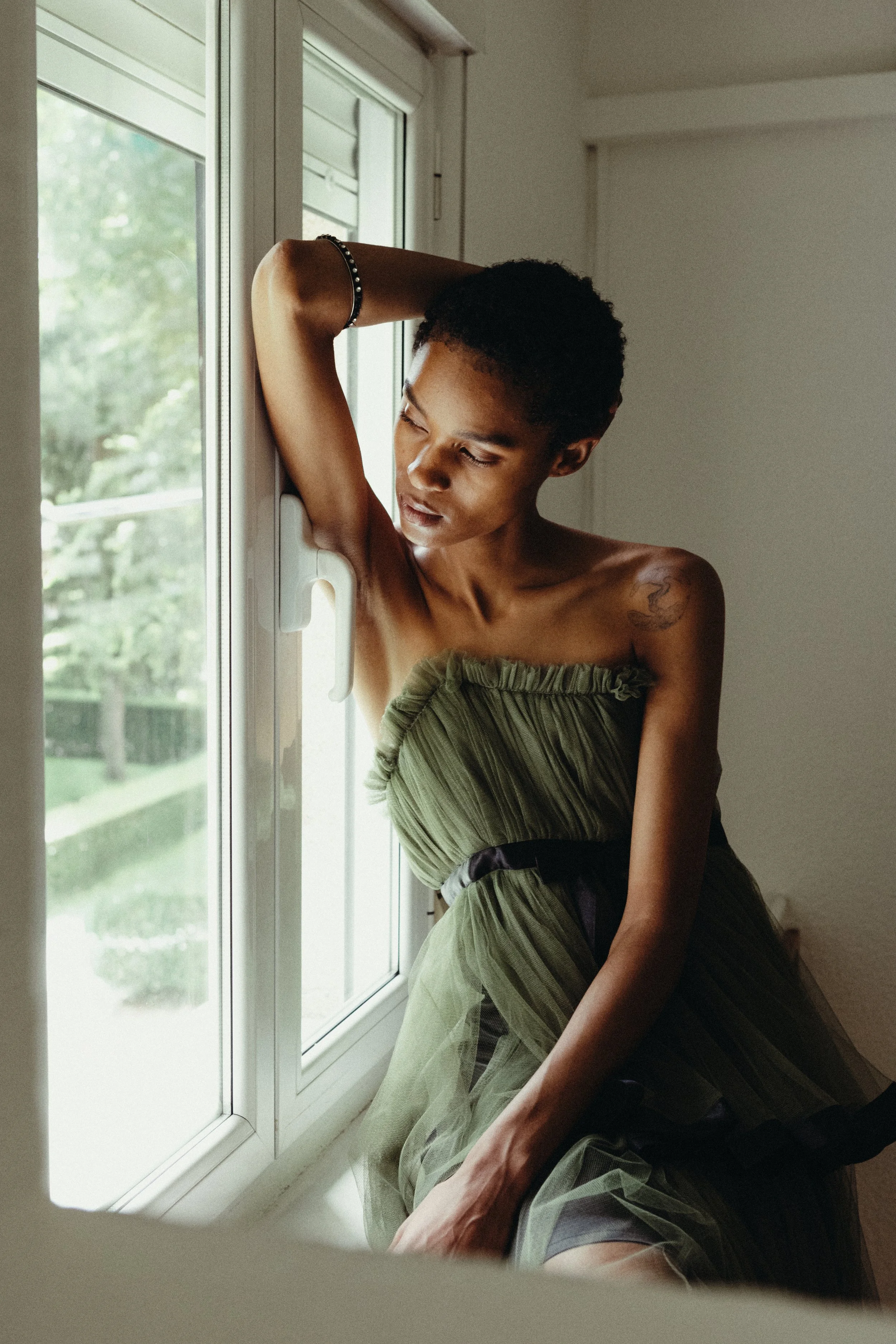 A woman with short curly hair and a tattoo on her shoulder is sitting by a window, wearing a green dress, with her eyes closed and her hand resting on her head.