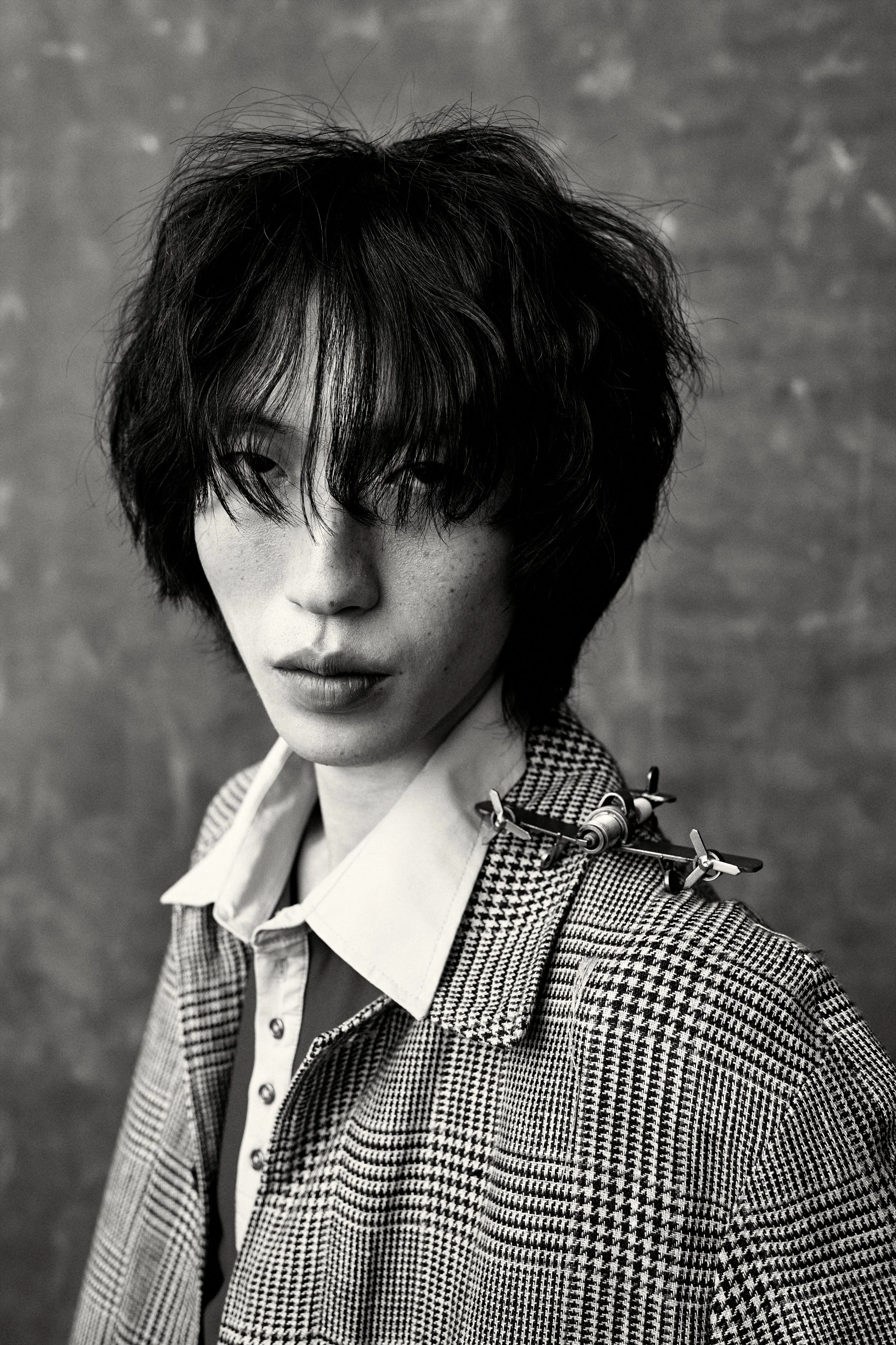 Black and white photograph of a young person with curly black hair, a checked blazer, and a patterned accessory on the shoulder, looking slightly downward with part of their face visible.
