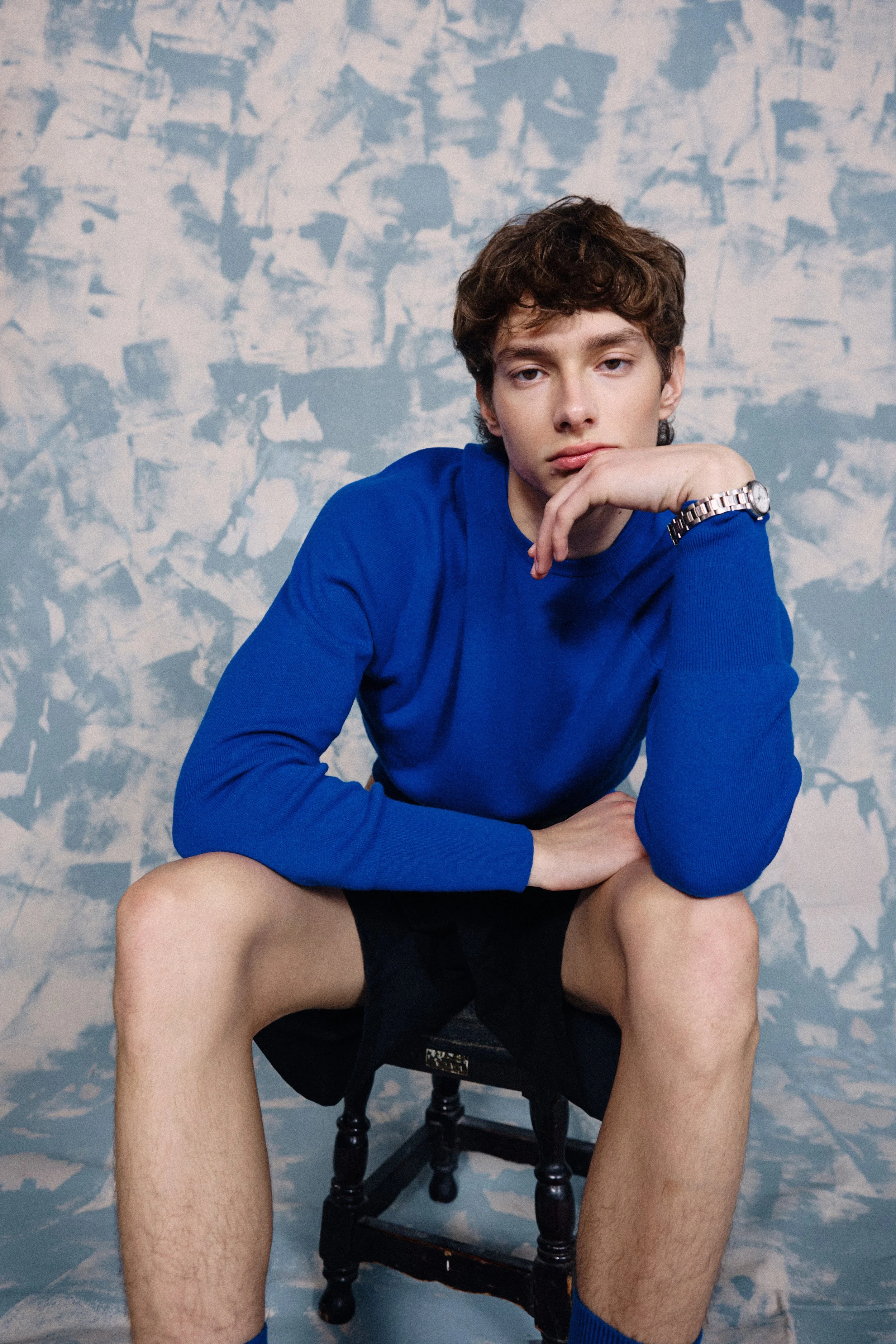 Young man sitting shirtless on a chair, wearing a blue long-sleeve shirt and a wristwatch, with a contemplative expression, against a abstract blue and white patterned wall background.