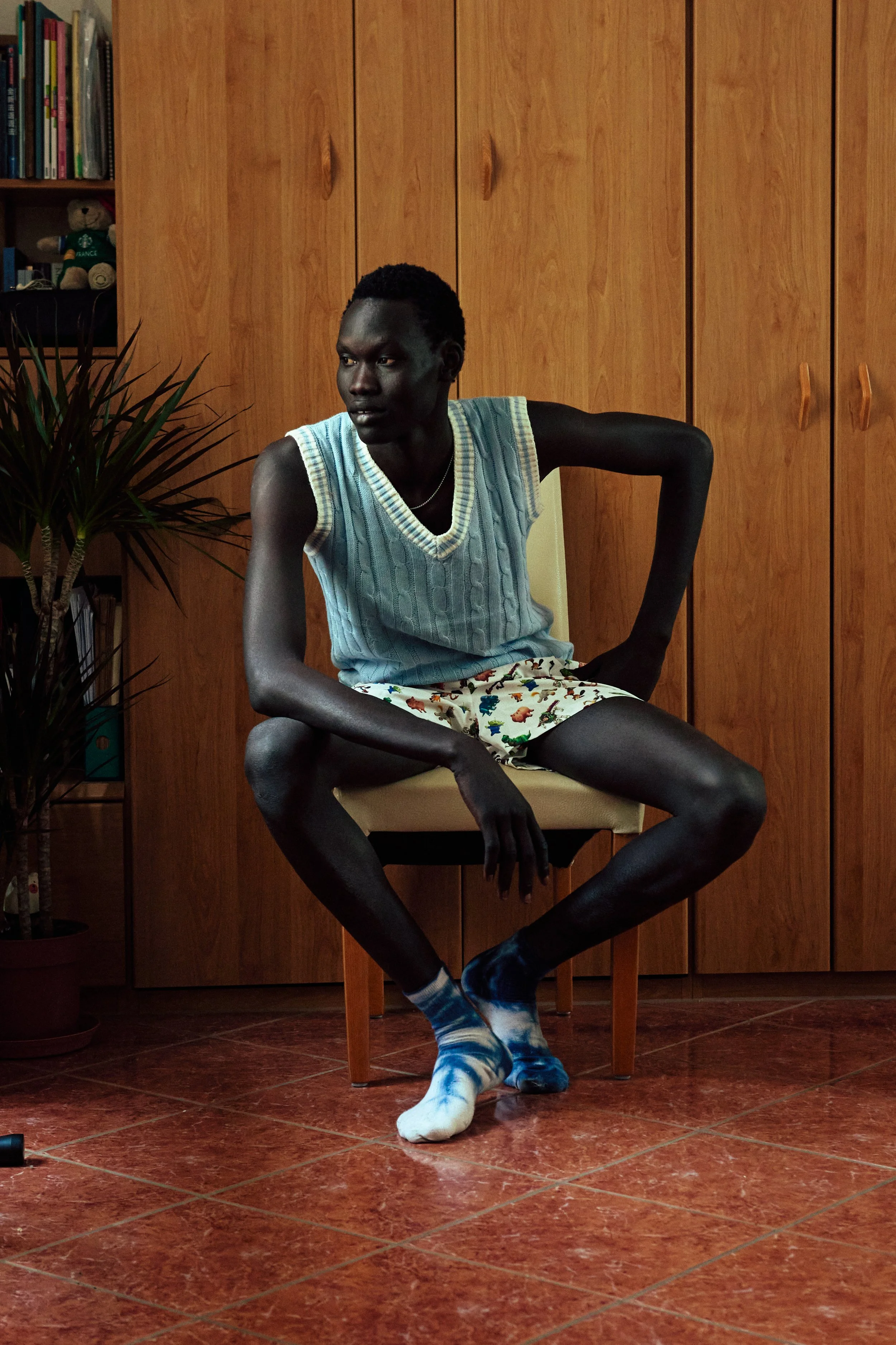 A young man with dark skin and short curly hair is sitting on a wooden chair in a room with wooden cupboards and a bookshelf. He is wearing a light blue knitted vest and patterned pajama shorts, with socks and shoes on. He has a serious expression an