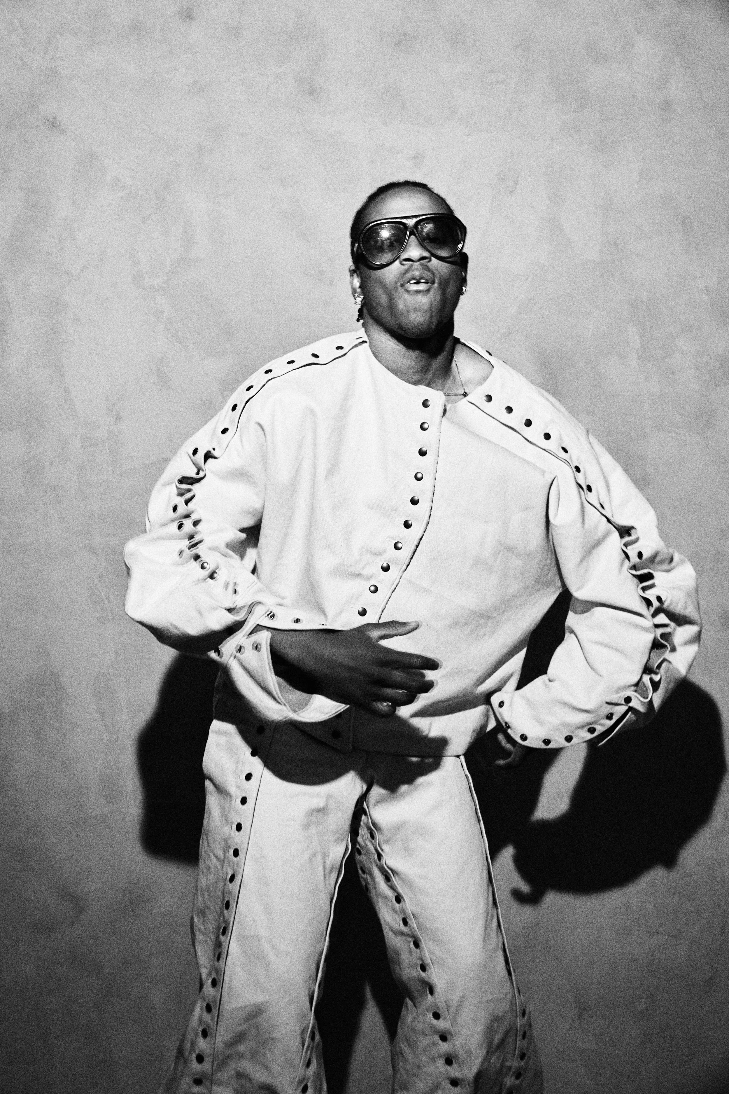 A person wearing oversized sunglasses and a white tracksuit with black buttons posing with one hand on their hip against a plain wall. The image is in black and white.