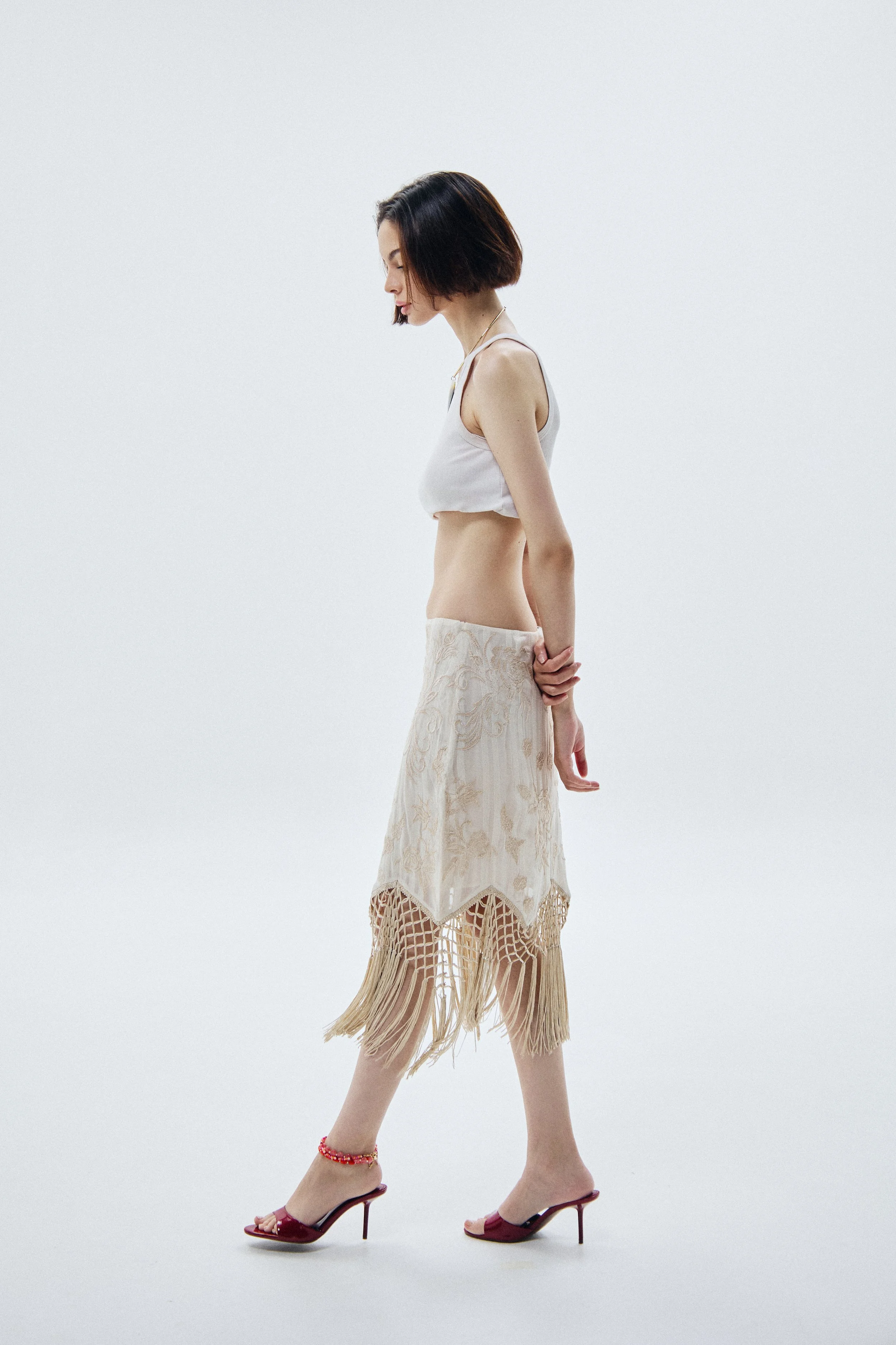 A woman with short dark hair posing in a white tank top and a long lace skirt with fringe, wearing red high-heeled sandals and a red ankle bracelet, standing against a plain white background.
