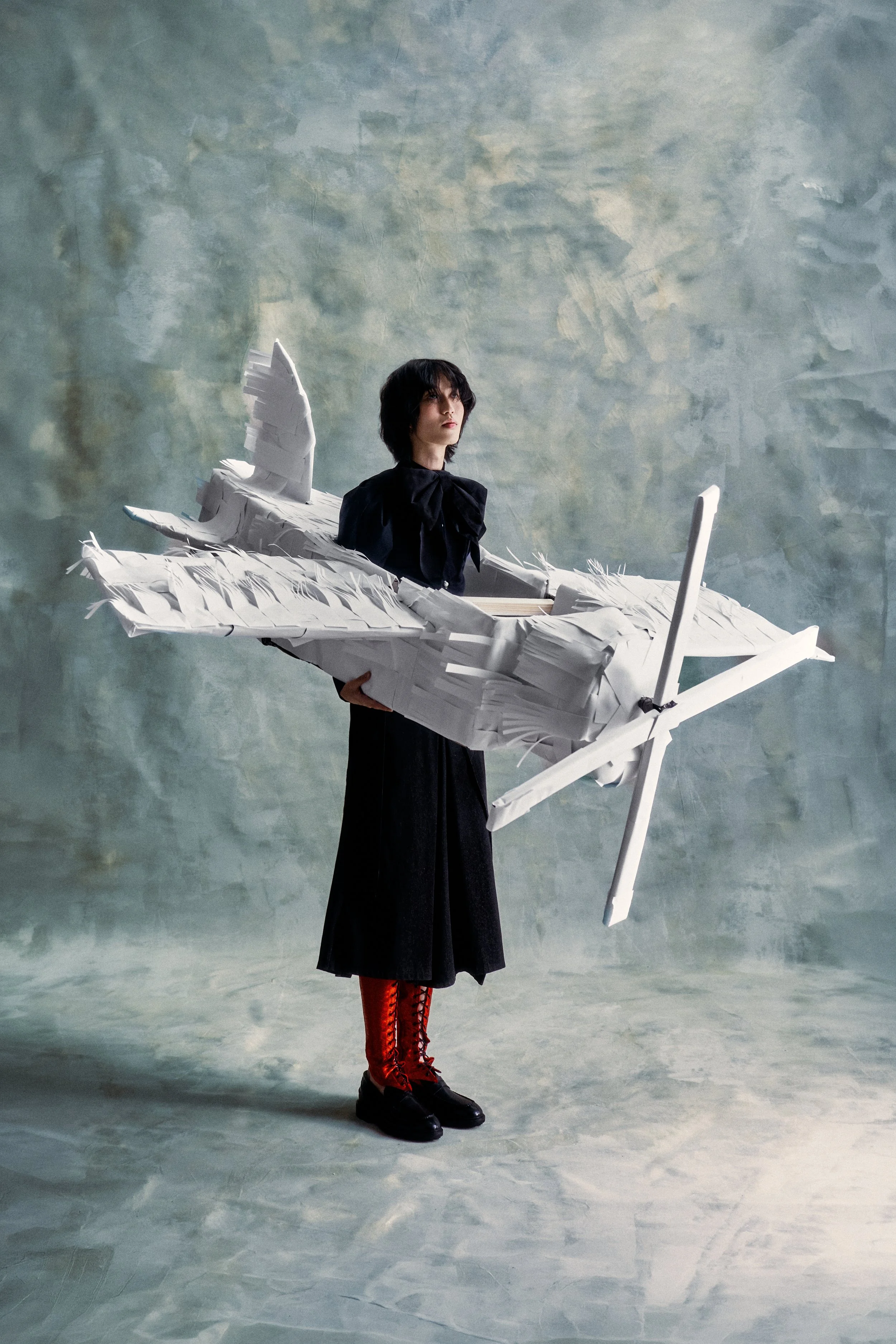 Person with black hair, dressed in black with red boots, holding a large paper airplane sculpture, standing on a textured grey background.