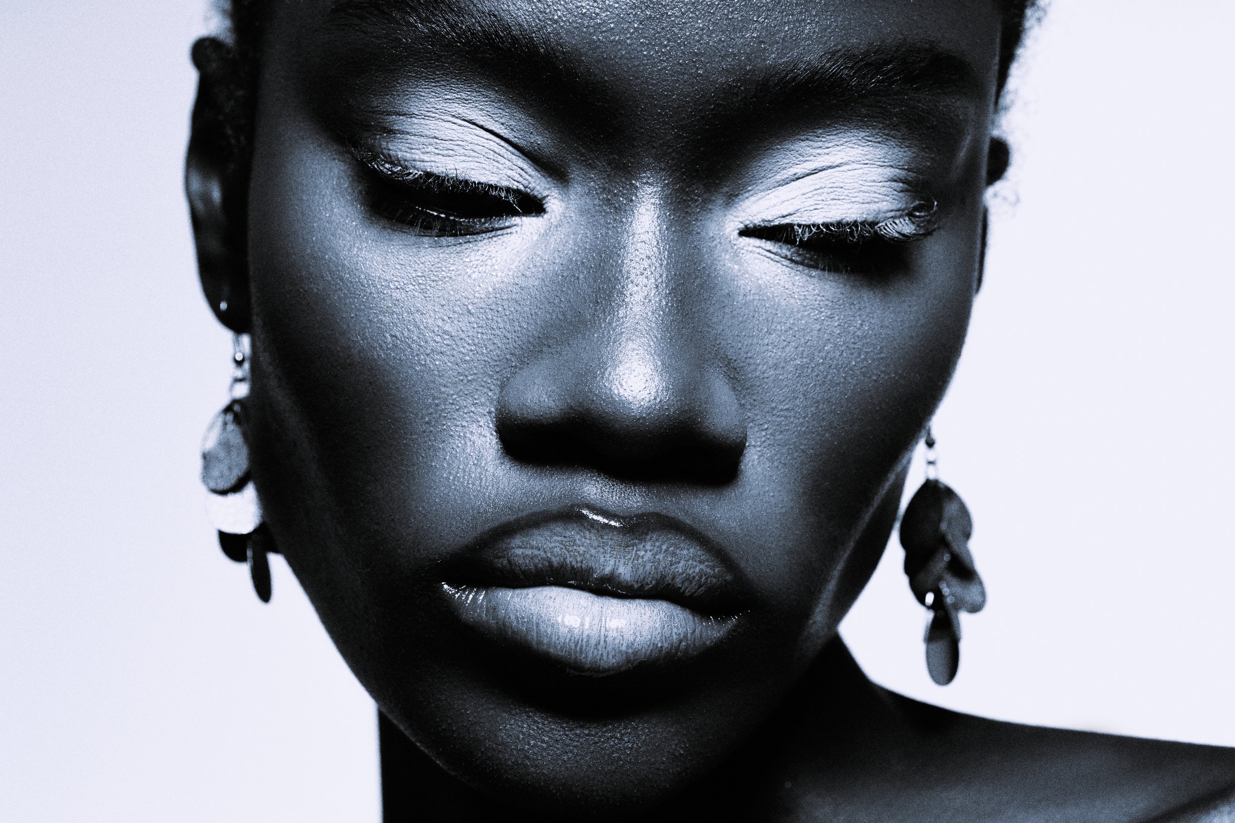 Close-up of a black woman's face with eyes closed, wearing earrings, with a neutral background.