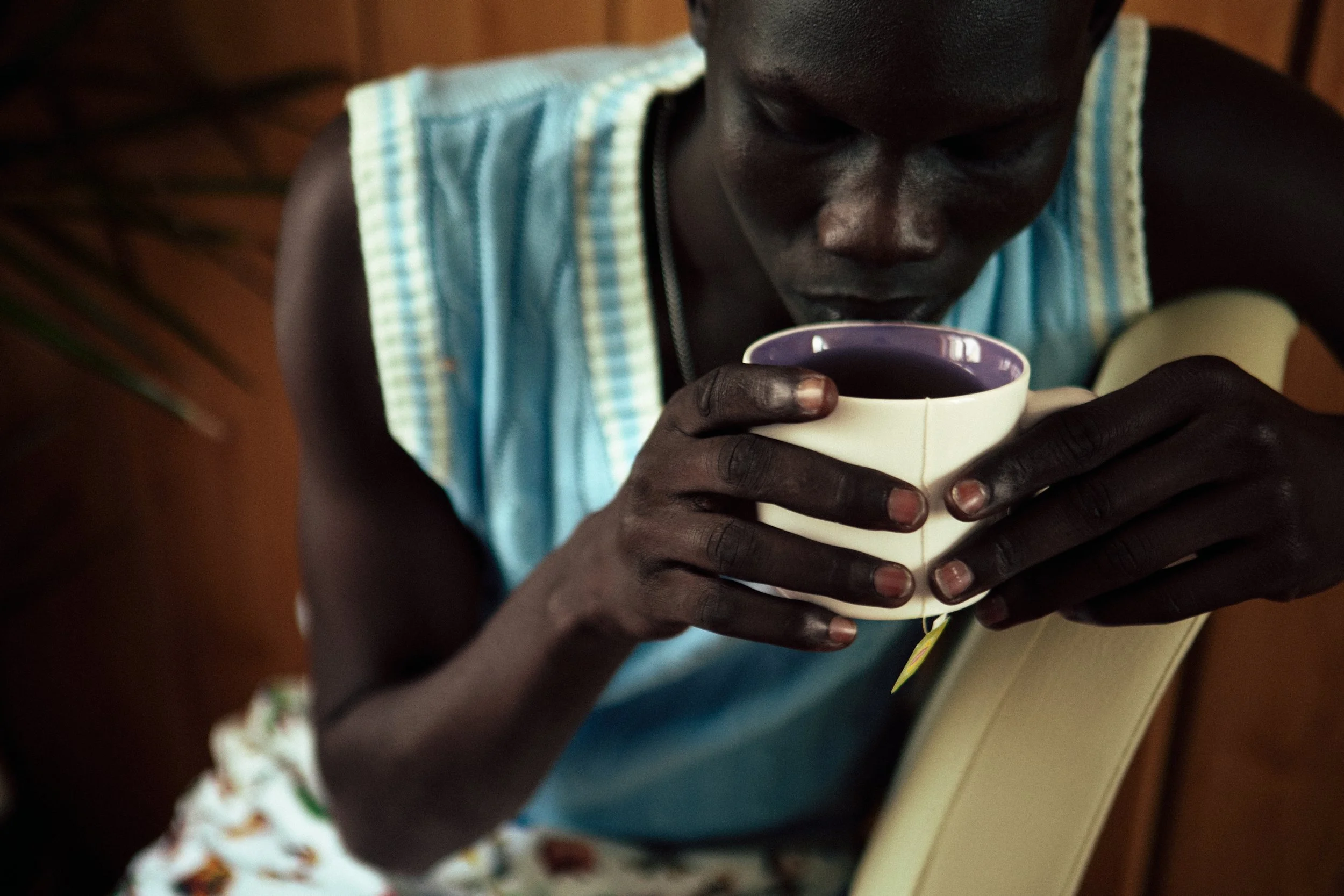 A person with dark skin holding a white mug with a purple interior, drinking from it. The person is wearing a sleeveless shirt with light blue, yellow, and white patterns and is sitting at a table with a patterned cloth.