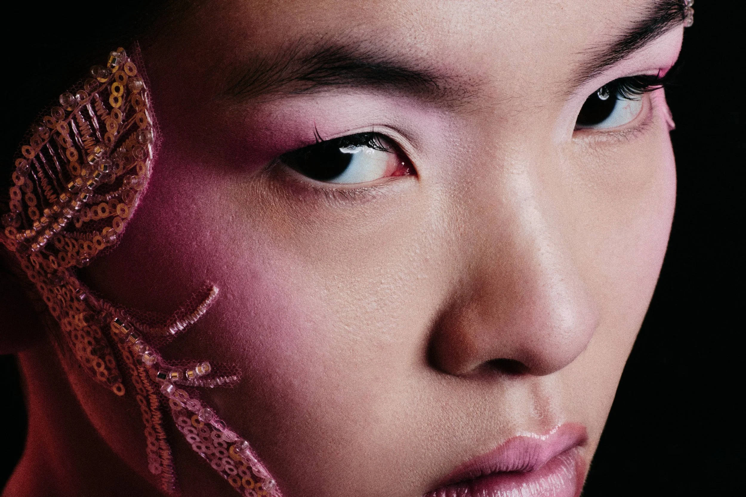 Close-up of a woman's face with pink eye makeup, black eyeliner, and long dark eyelashes. She is wearing a headscarf decorated with pink sequins and beads.