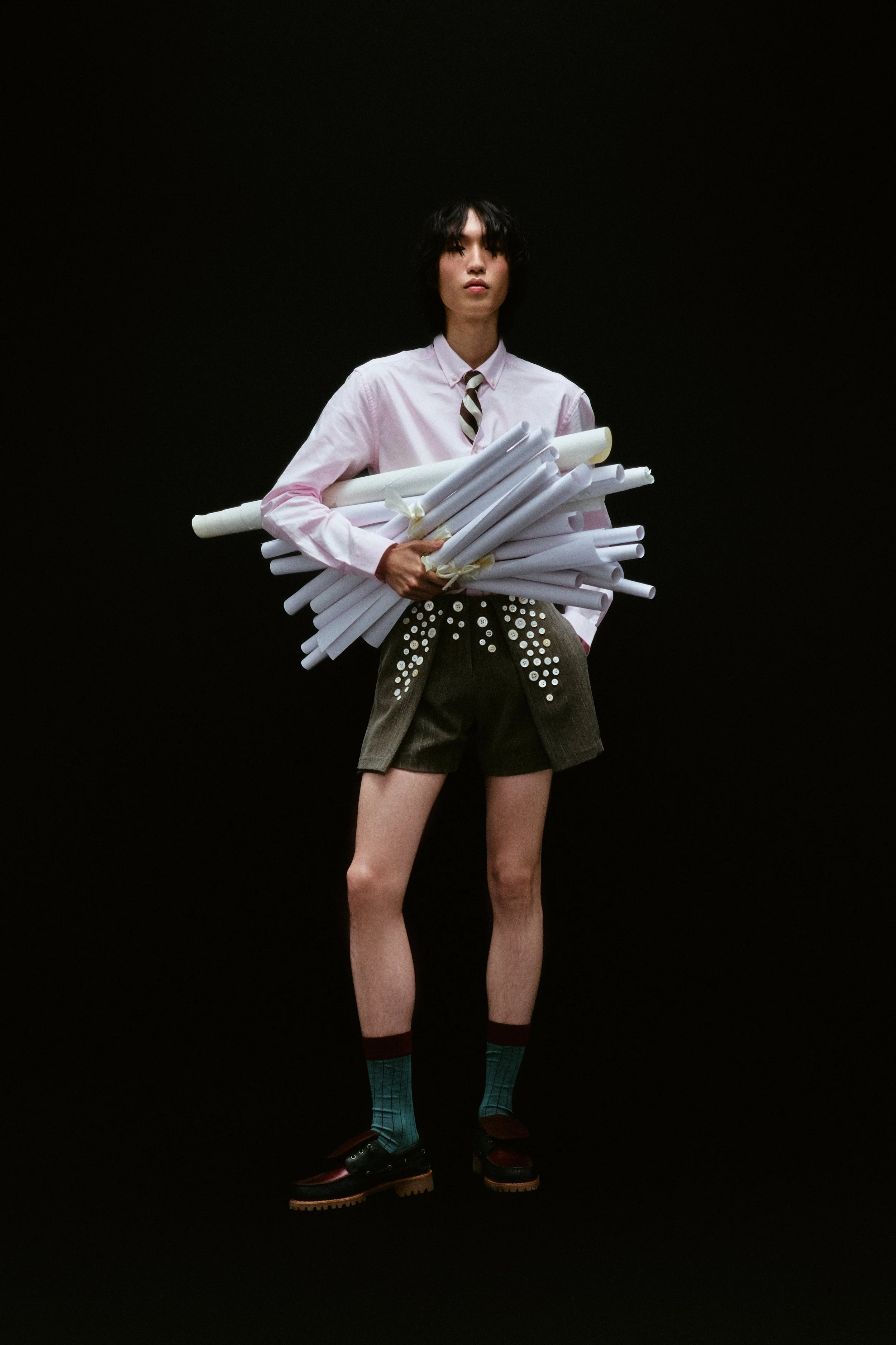 Person standing against a black background, wearing a pink shirt, a patterned tie, shorts with decorative buttons, colorful socks, and chunky shoes, holding rolled-up papers.