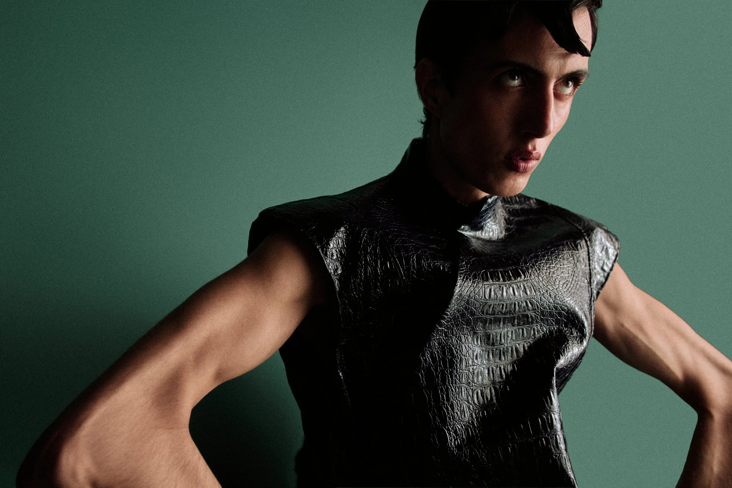 Close-up of a woman with short dark hair, intense gaze, and wearing a textured black sleeveless top against a plain green background.