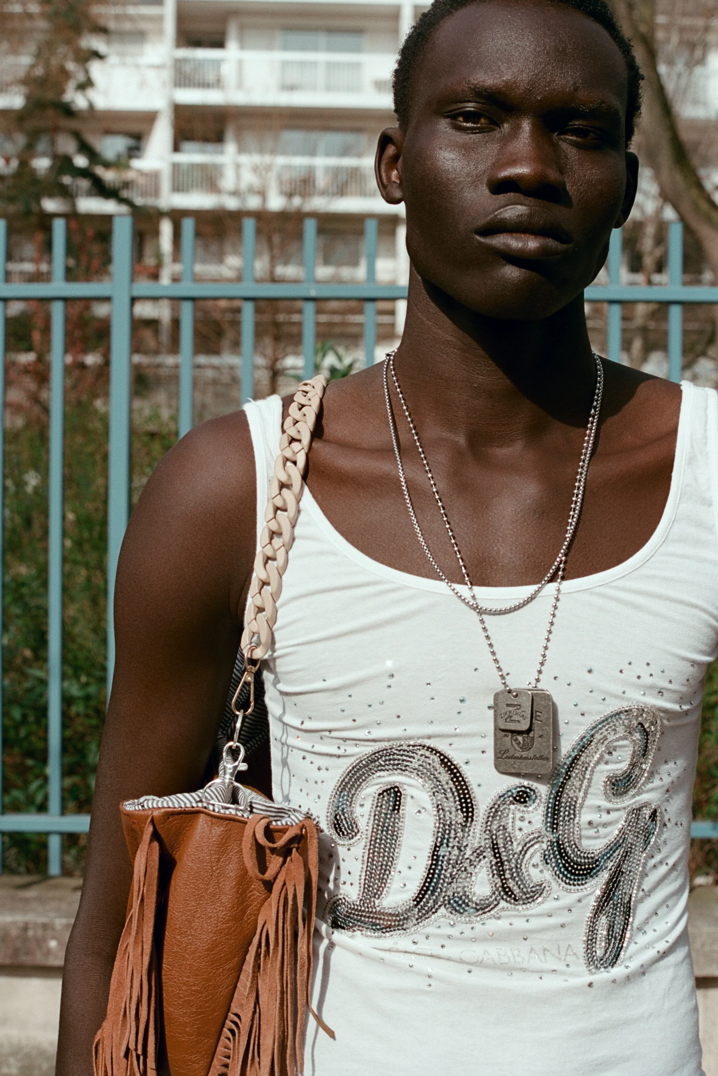 Close-up of a woman with dark skin and short hair wearing a white tank top with glittering designs, layered silver necklaces, a large beige chain strap bag with fringes, and a dog tag necklace. She is standing outdoors in front of a blue metal fence 
