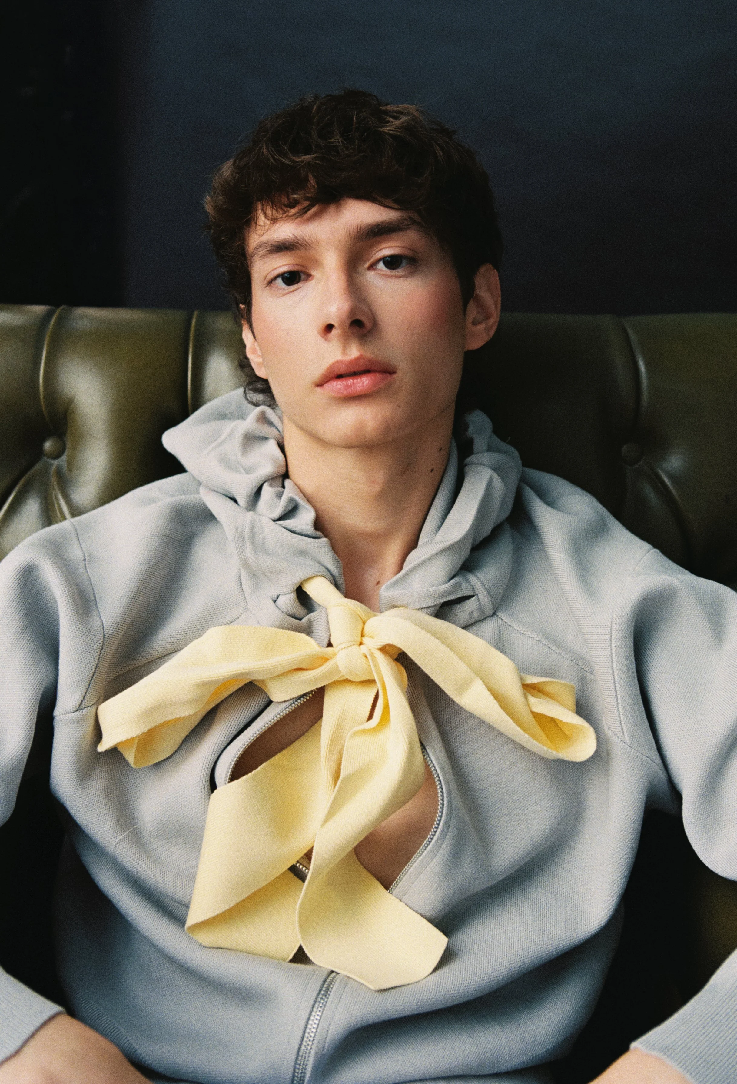 A young man with dark curly hair and fair skin sits on a dark green leather chair, wearing a light gray hoodie with a yellow bow around his neck.