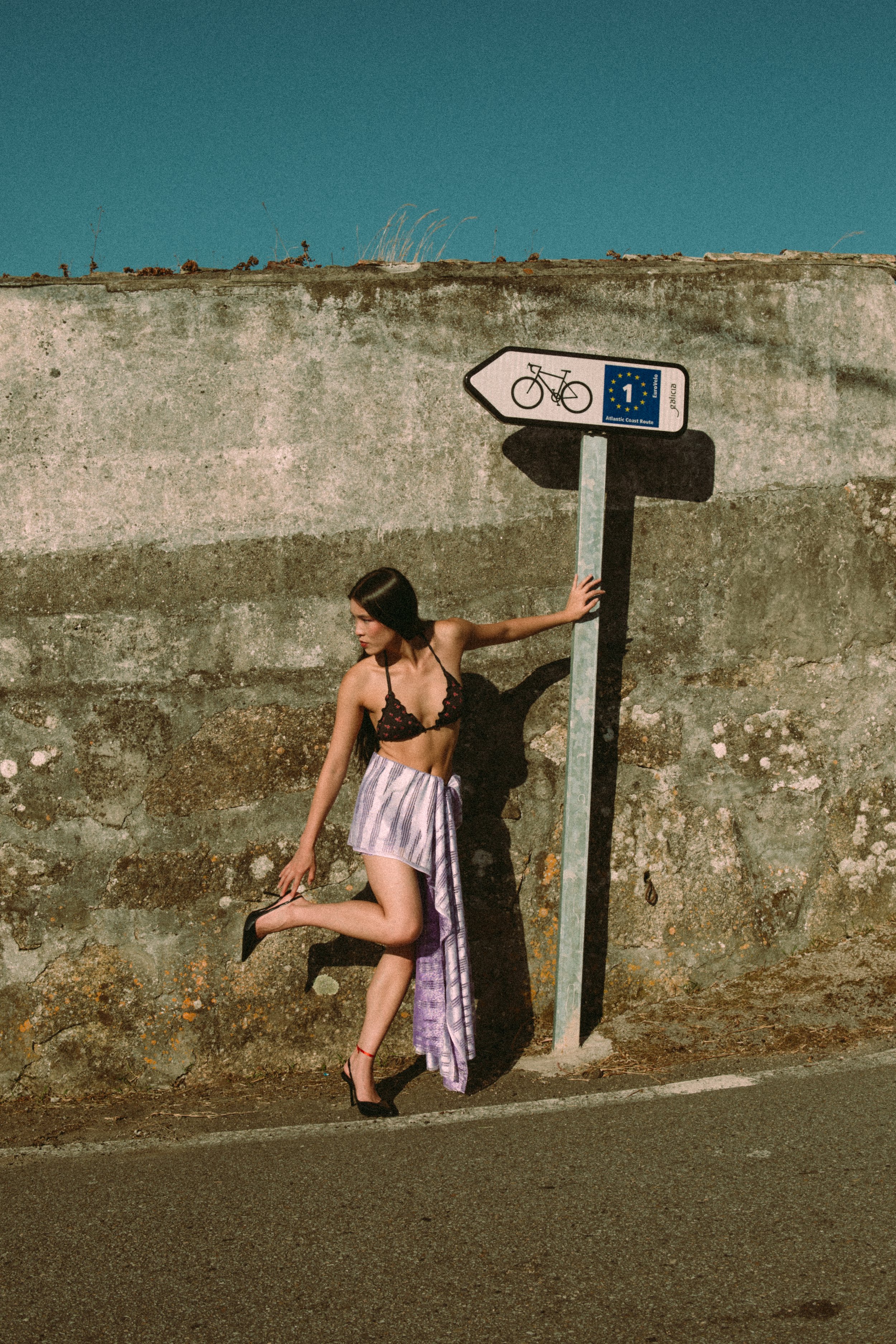 A young woman in high heels, a black bra with red accents, and a long purple skirt posing by a concrete wall. She is holding a bicycle sign with her left hand, with her right hand touching the ground, and one foot on the ground while the other leg is