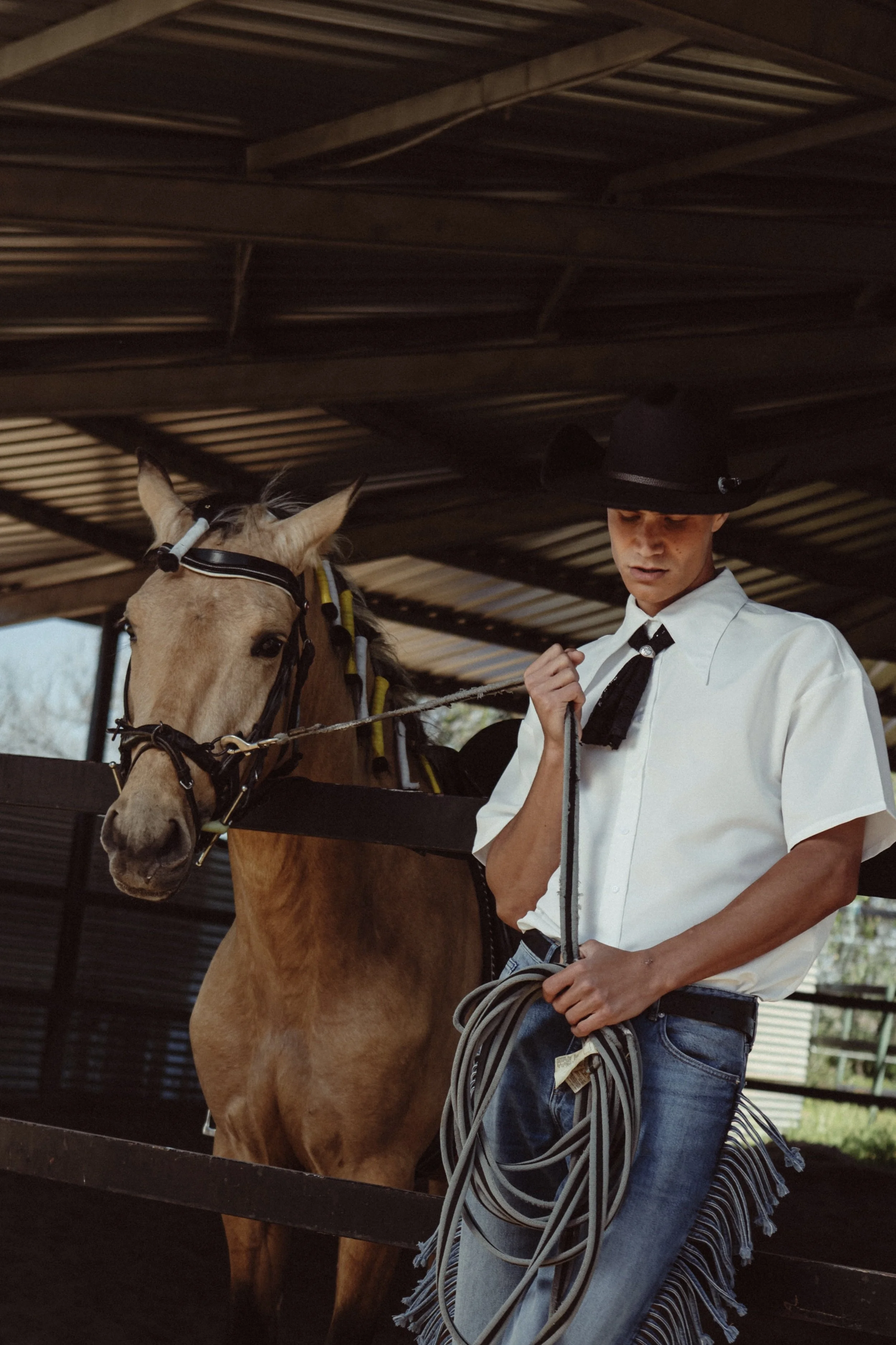 A young man in a white western shirt, wearing a black cowboy hat, holding a lasso rope, standing next to a light brown horse with a bridle, inside a wooden barn.