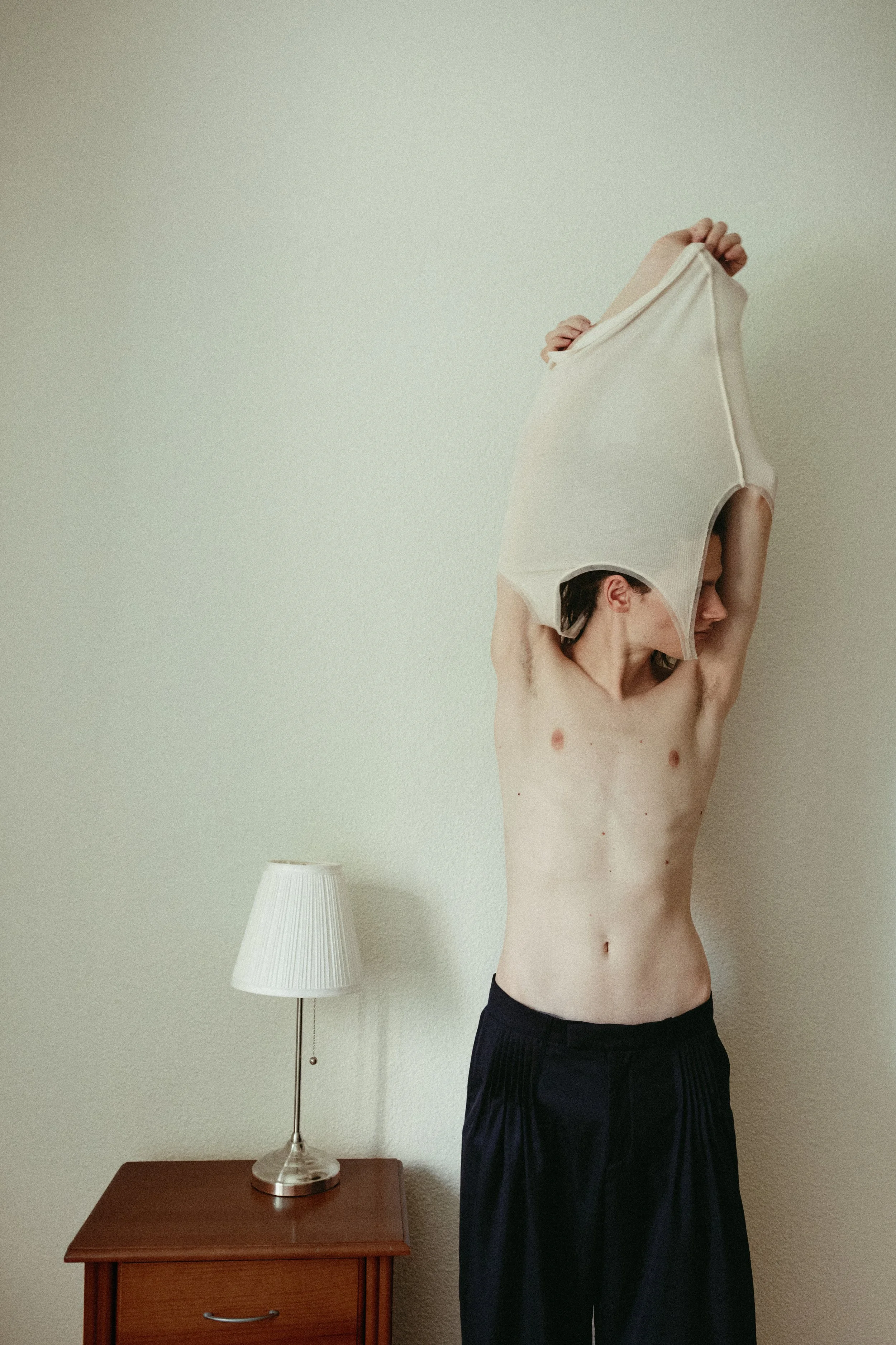 A person is standing without a shirt, pulling a white T-shirt over their head in a room with beige walls. A wooden bedside table with a drawer and a white lampshade is located to the left.