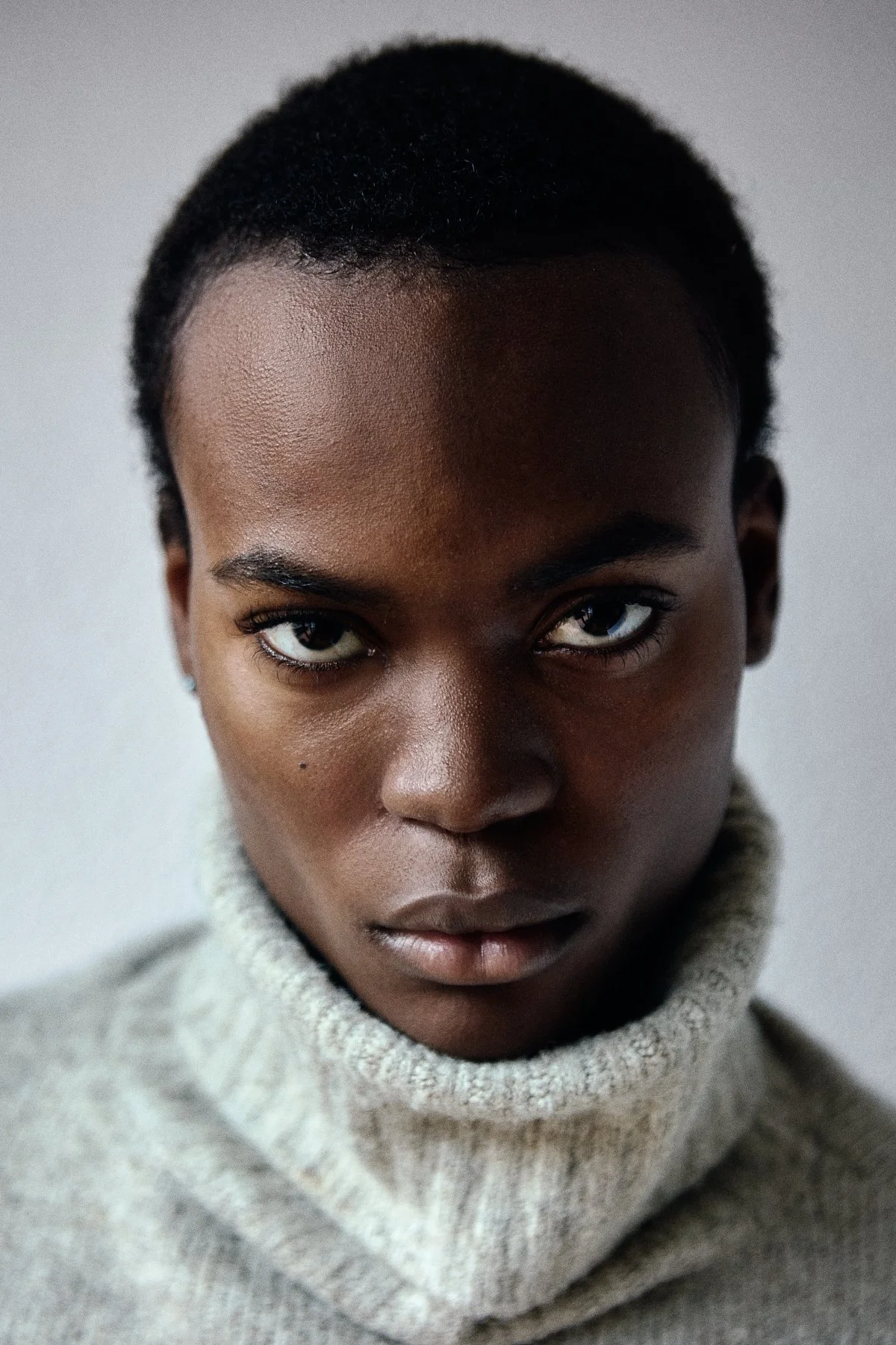 Close-up portrait of a young African American woman with intense gaze, short curly hair, and wearing a light gray turtleneck sweater.