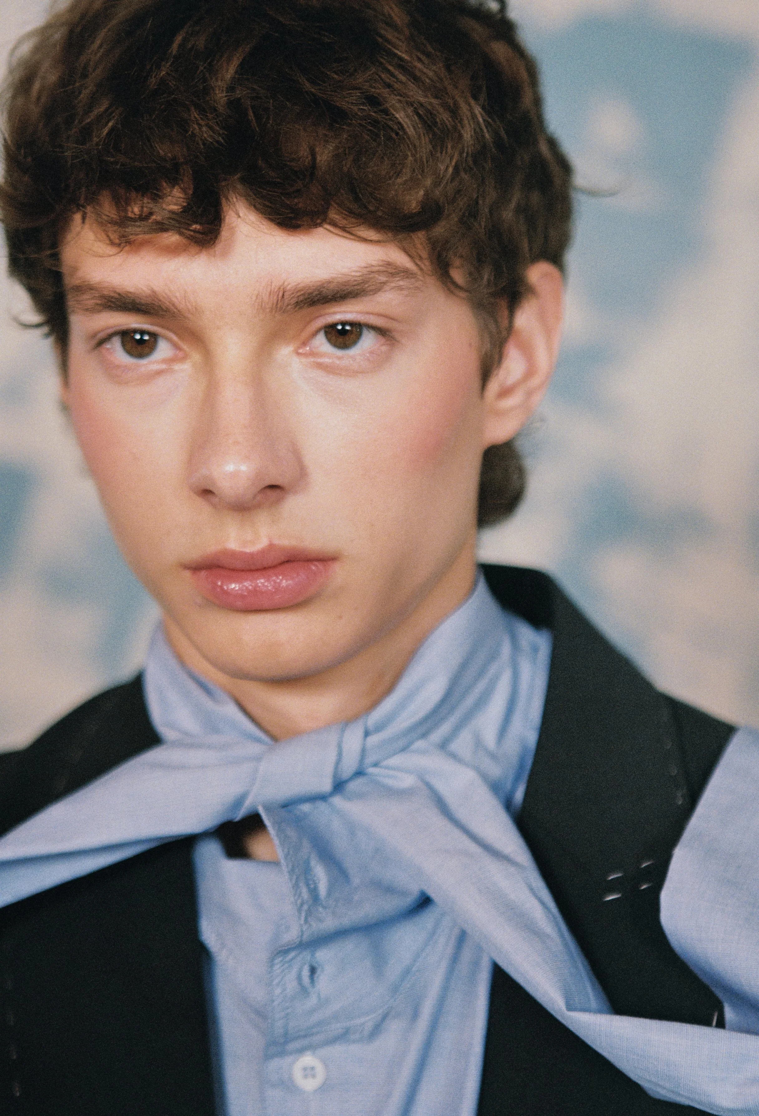 Close-up of a woman with short curly brown hair, wearing a black blazer and a light blue button-up shirt with a matching bow tie, against a cloudy sky background.
