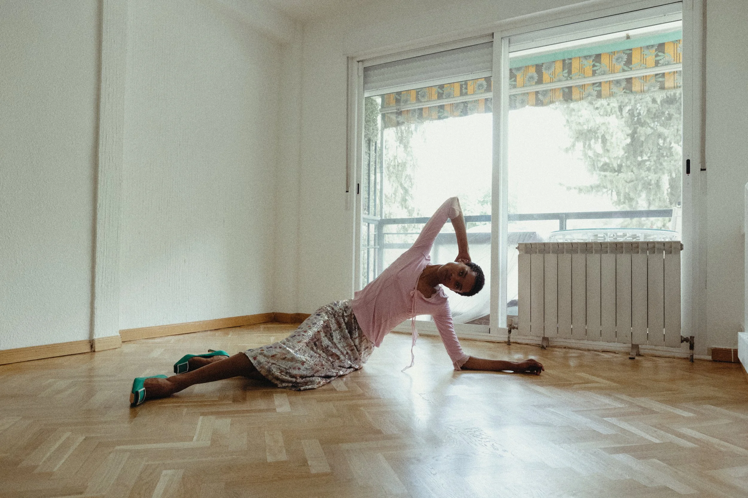 A woman in a pink top and floral skirt doing a side plank yoga pose on a wooden floor in a room with large windows.