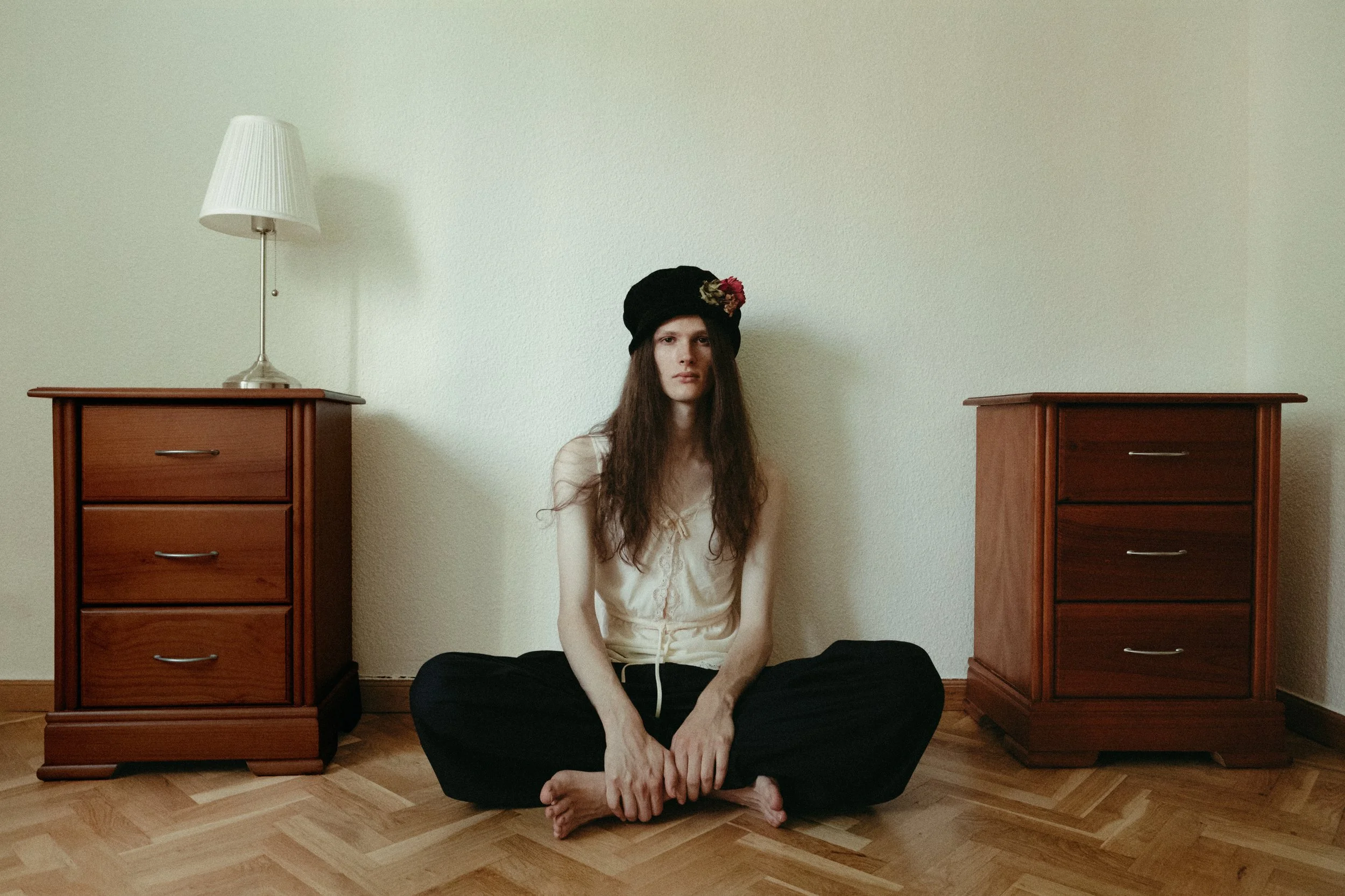 A woman with long brown hair, wearing a black hat with flowers, a cream-colored top, and black pants, sitting cross-legged on a wooden floor between two wooden dressers in a room with plain light-colored walls.