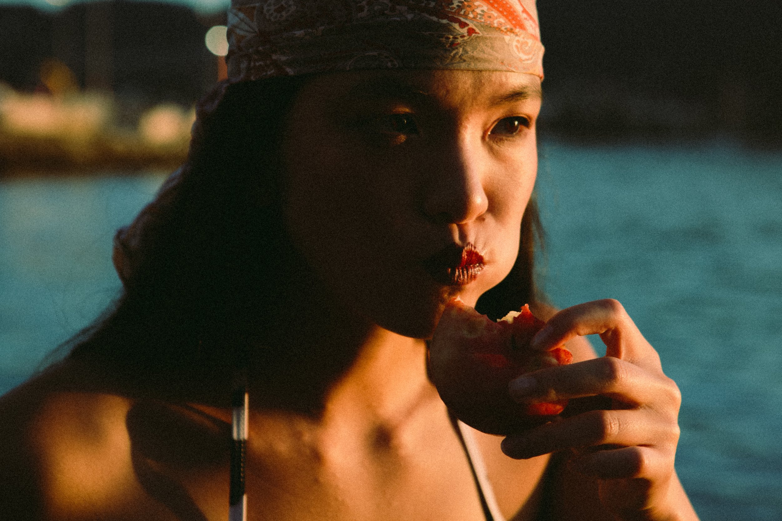A woman with tan skin wearing a pink patterned headscarf and thin black straps is eating a slice of pizza at night, with city lights and water blurred in the background.
