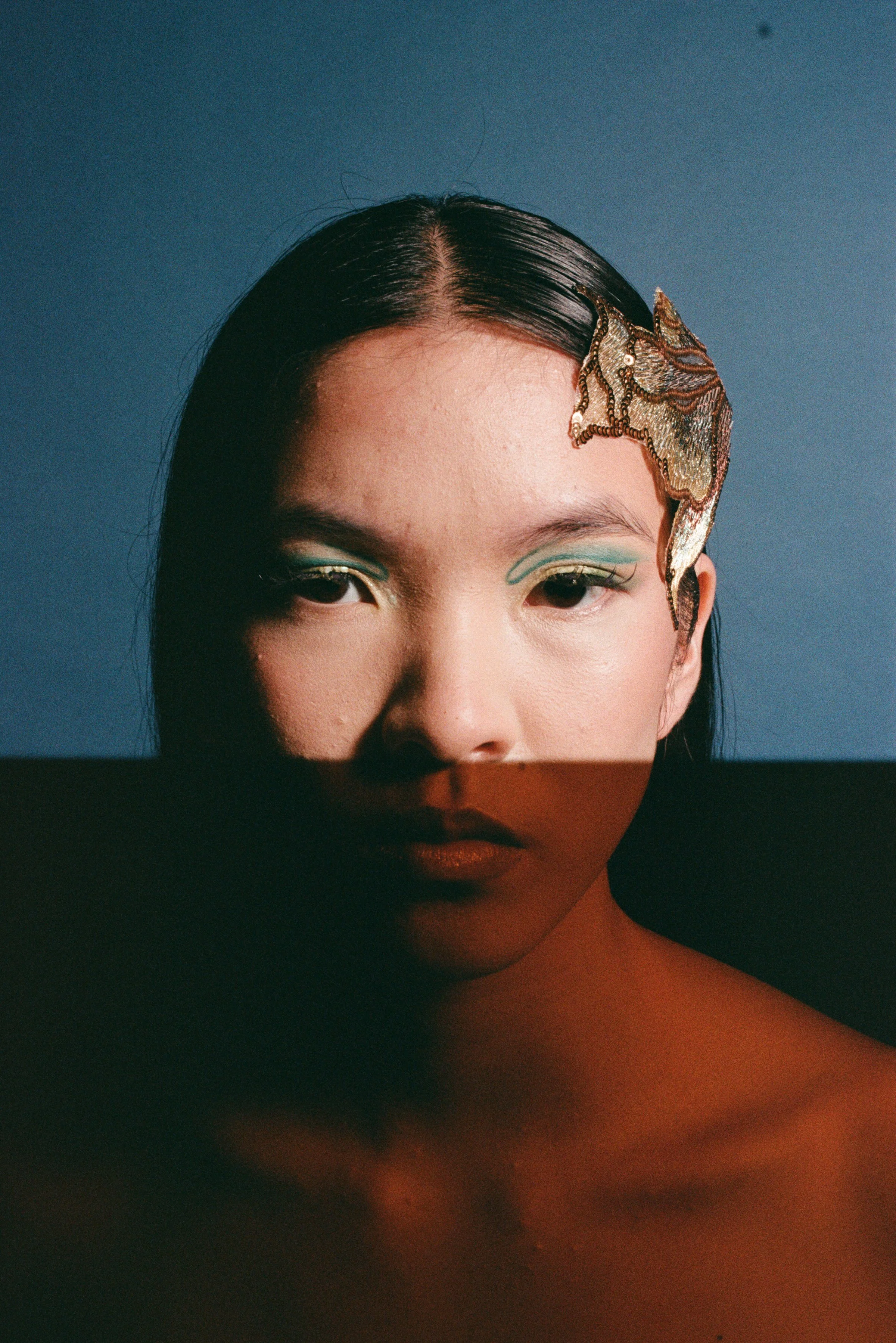 Close-up portrait of a woman with black hair and artistic makeup, including teal eyeliner and a textured gold and bronze accessory on the top right of her forehead, set against a gradient blue background.