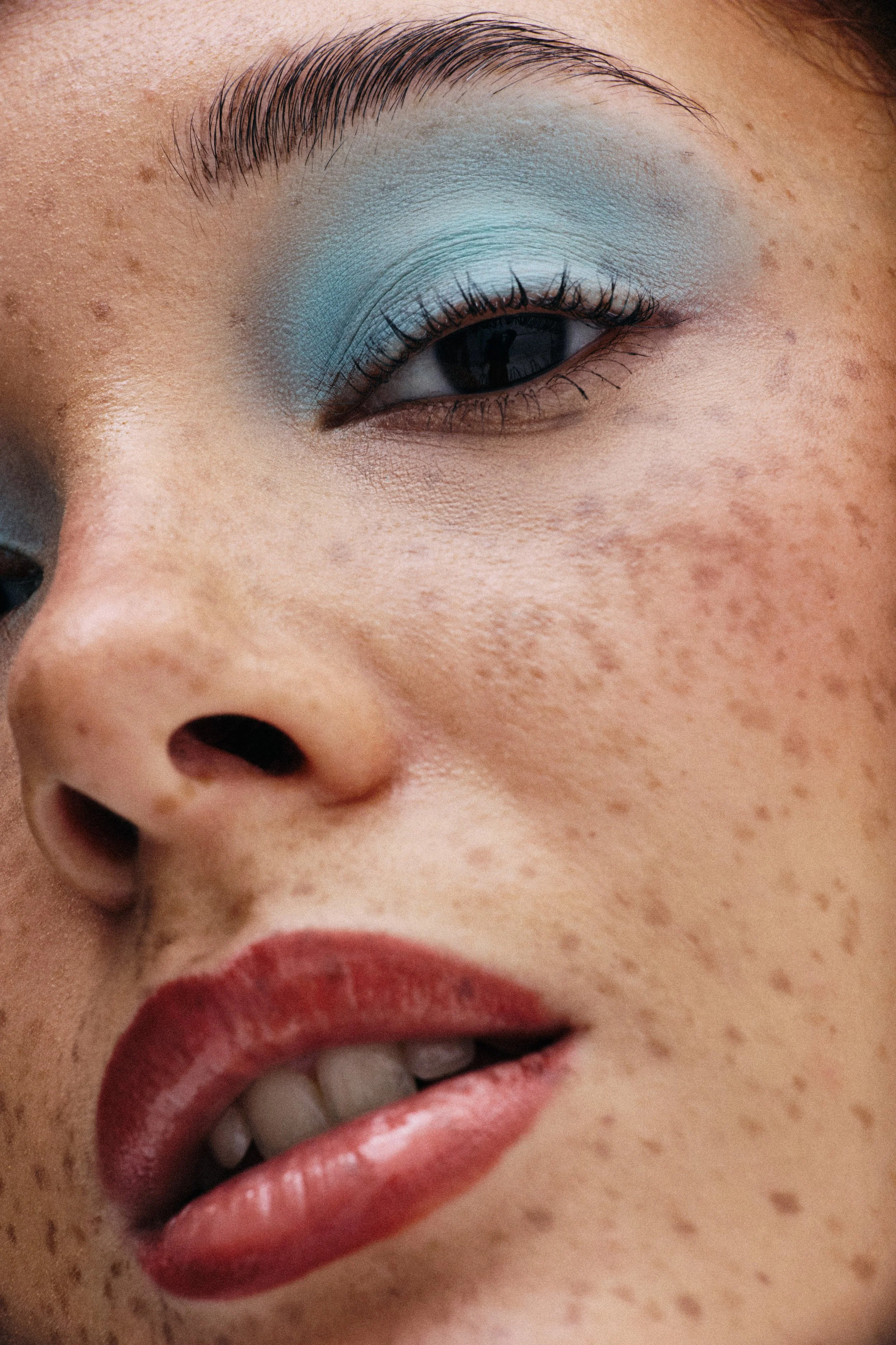 Close-up of a person's face showing a well-groomed eyebrow, eye with blue eyeshadow, and red lipstick. The person's skin has freckles.