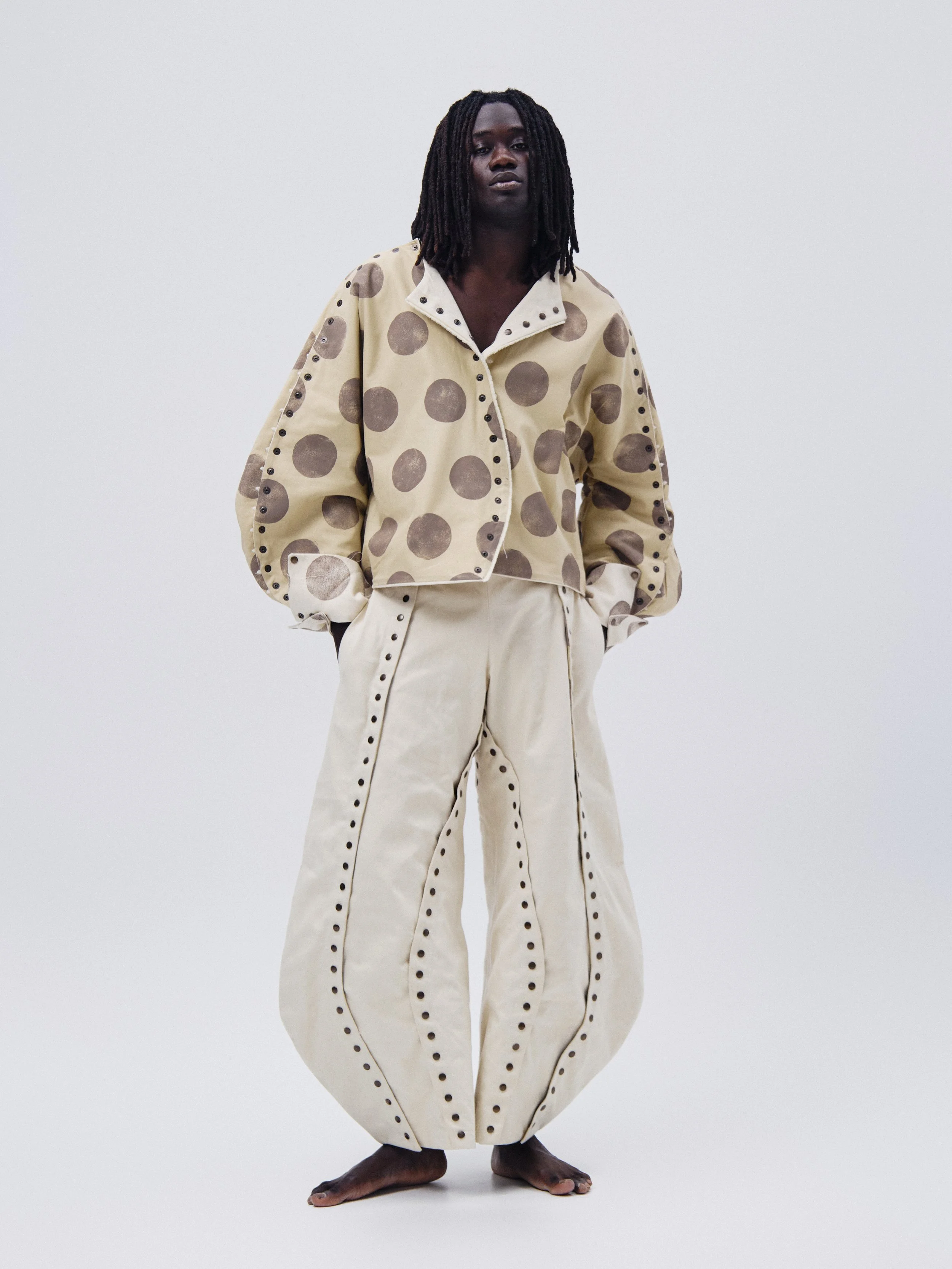 A person standing barefoot, wearing wide-leg cream-colored pants with black stitching and a beige jacket with dark brown polka dots, against a plain white background.