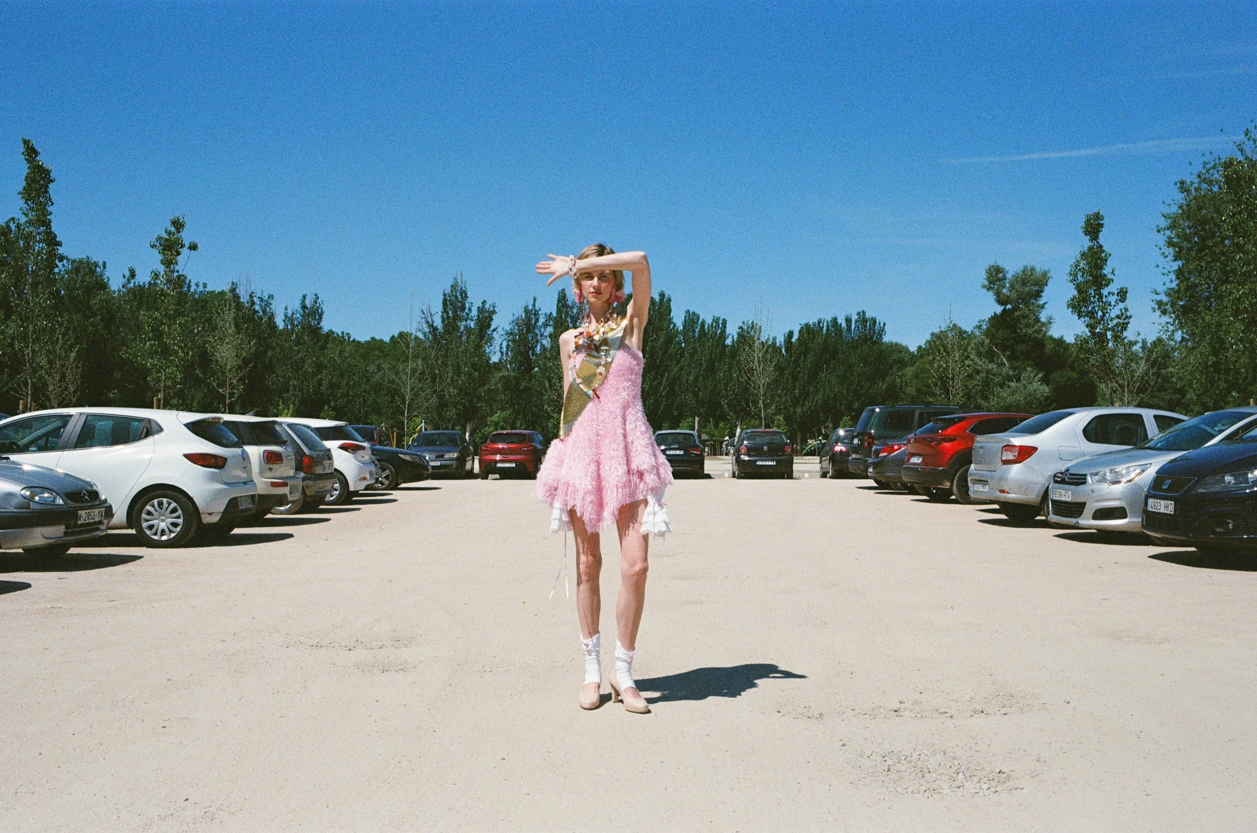 A woman standing in a parking lot, dressed in a pink, fuzzy dress with jewelry, wearing high heels, and shielding her eyes from the sun with her hand, with cars and trees under a clear blue sky.