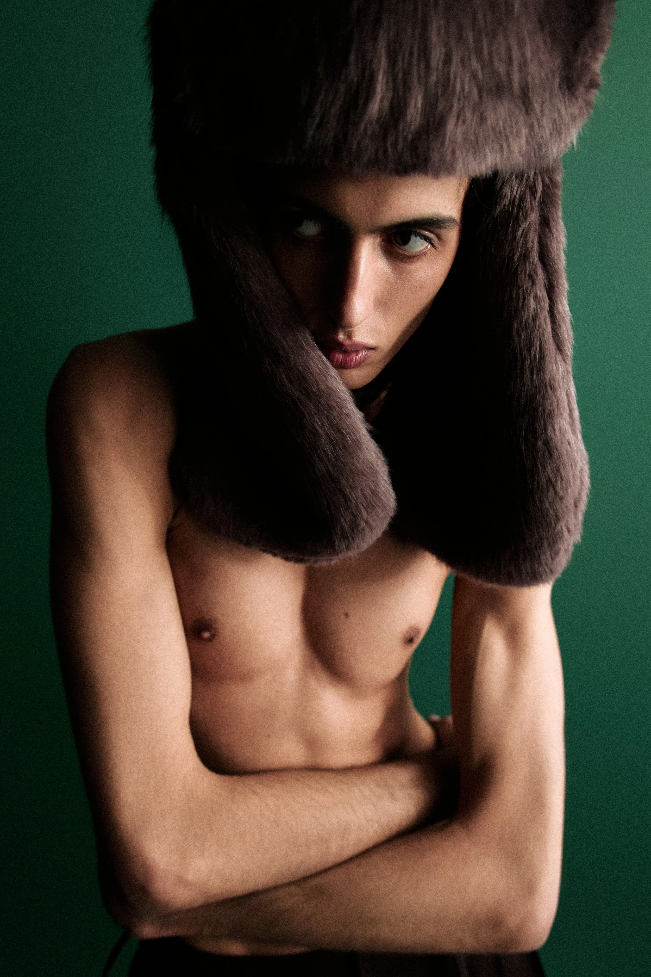 A partially nude person with short hair, wearing a brown fur hat and fur collar, standing against a green background, with arms crossed in front of their torso.