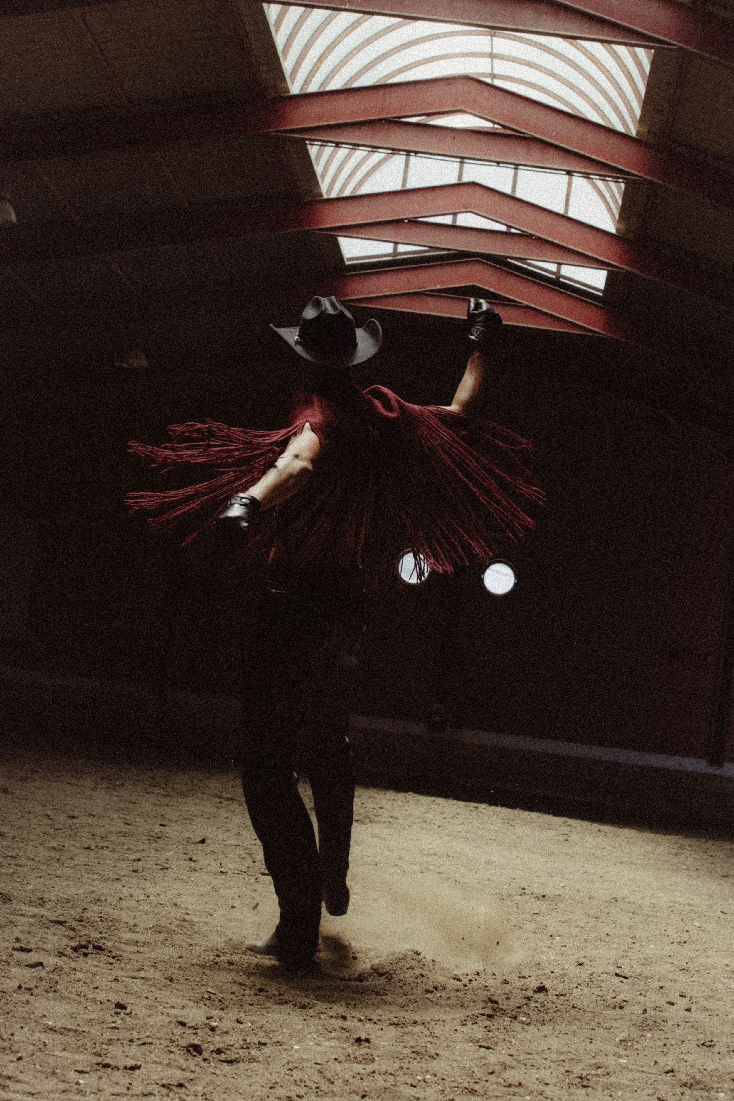 A person dressed as a cowboy with a hat, red fringed dress, and black boots performing a dance inside a building with a high, arching ceiling and a dirt floor.