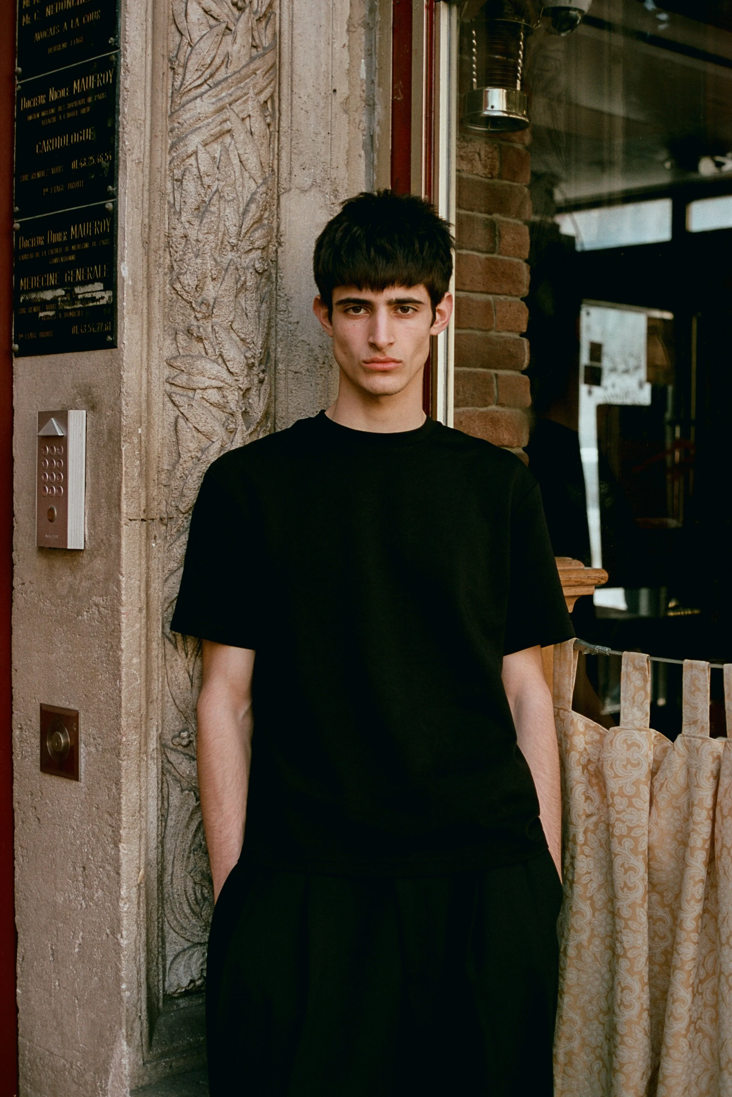 A young man with dark hair and a serious expression, wearing a black t-shirt, standing outside next to a building with textured stone and brick walls, near an intercom panel and a sign with medical office information.