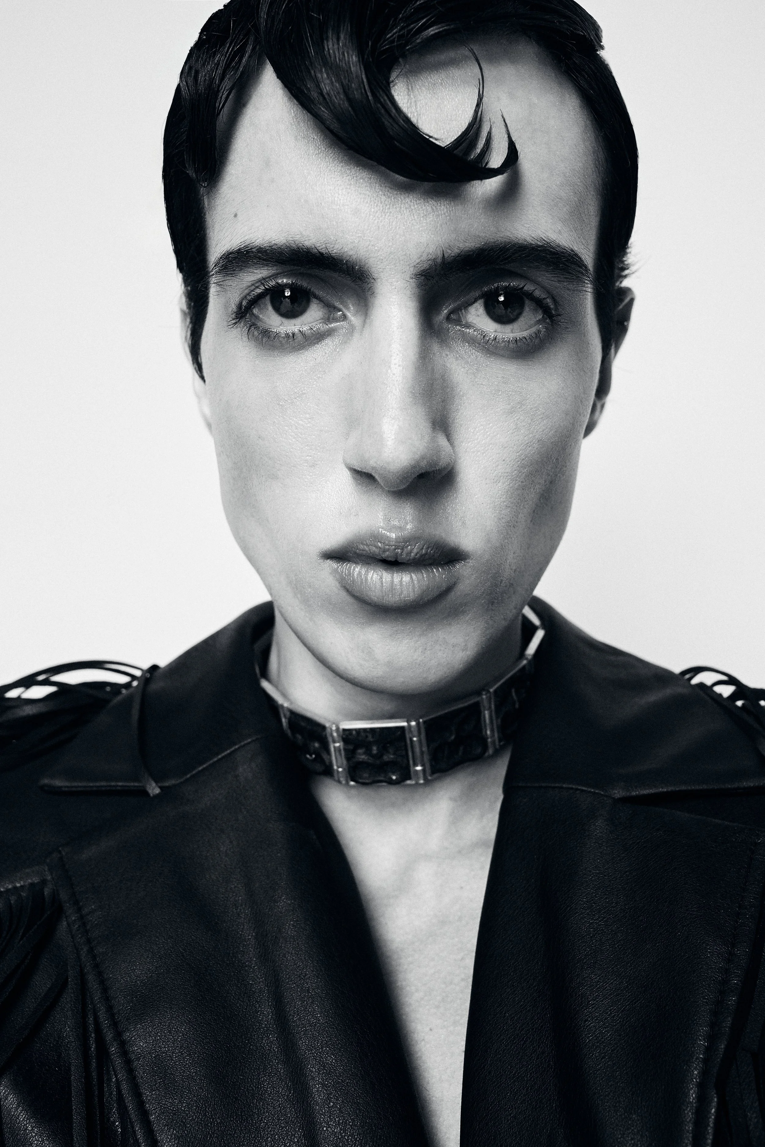 Black and white portrait of a woman with short, dark hair and piercing eyes, wearing a leather jacket and a chunky metallic choker.